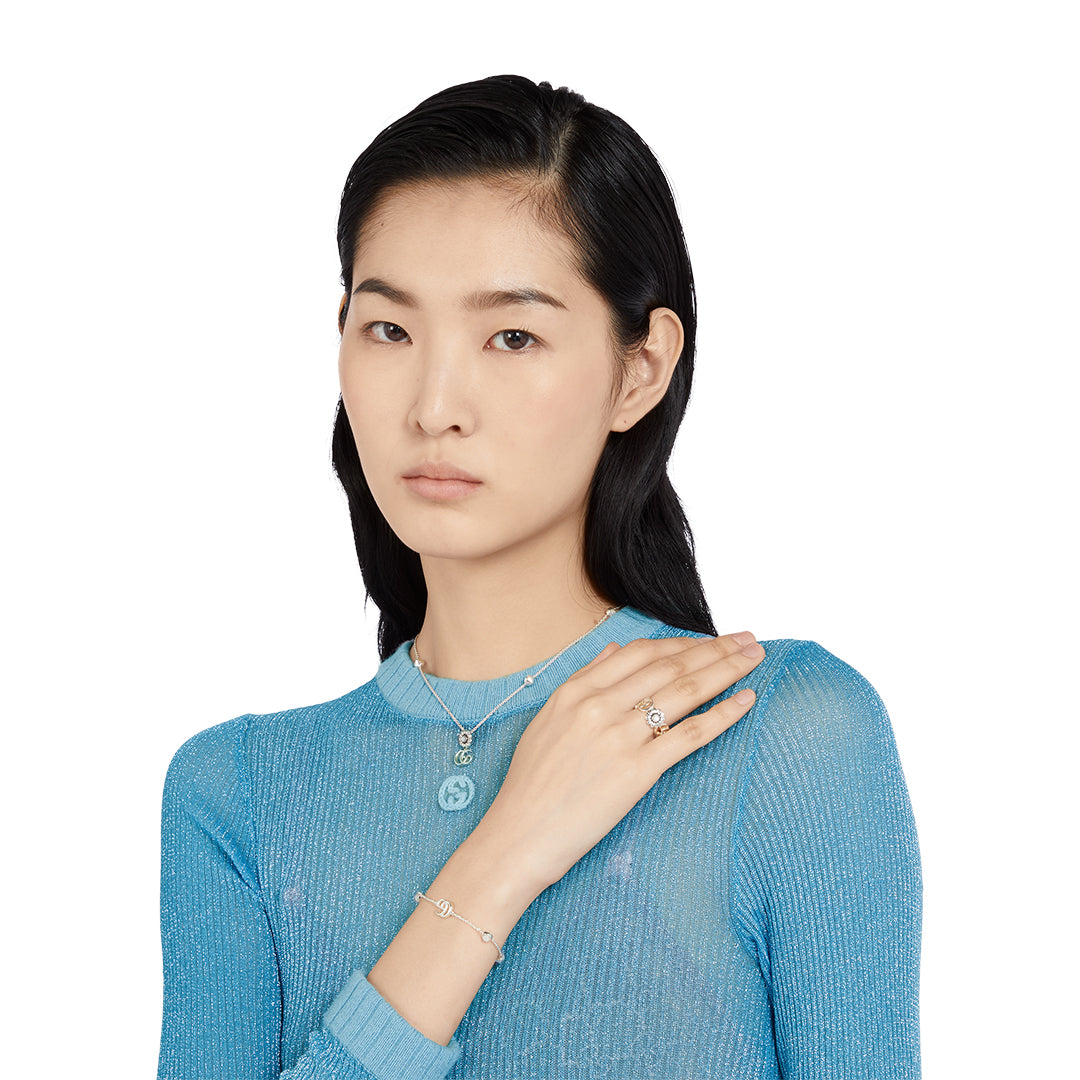 A person with long, dark hair in a sheer blue top poses against a white backdrop, wearing silver jewelry including a Gucci GG Marmont Sterling Silver Necklace Pendant by GUCCI Fine Jewellery, along with a mother-of-pearl accented bracelet and a ring on their left hand.