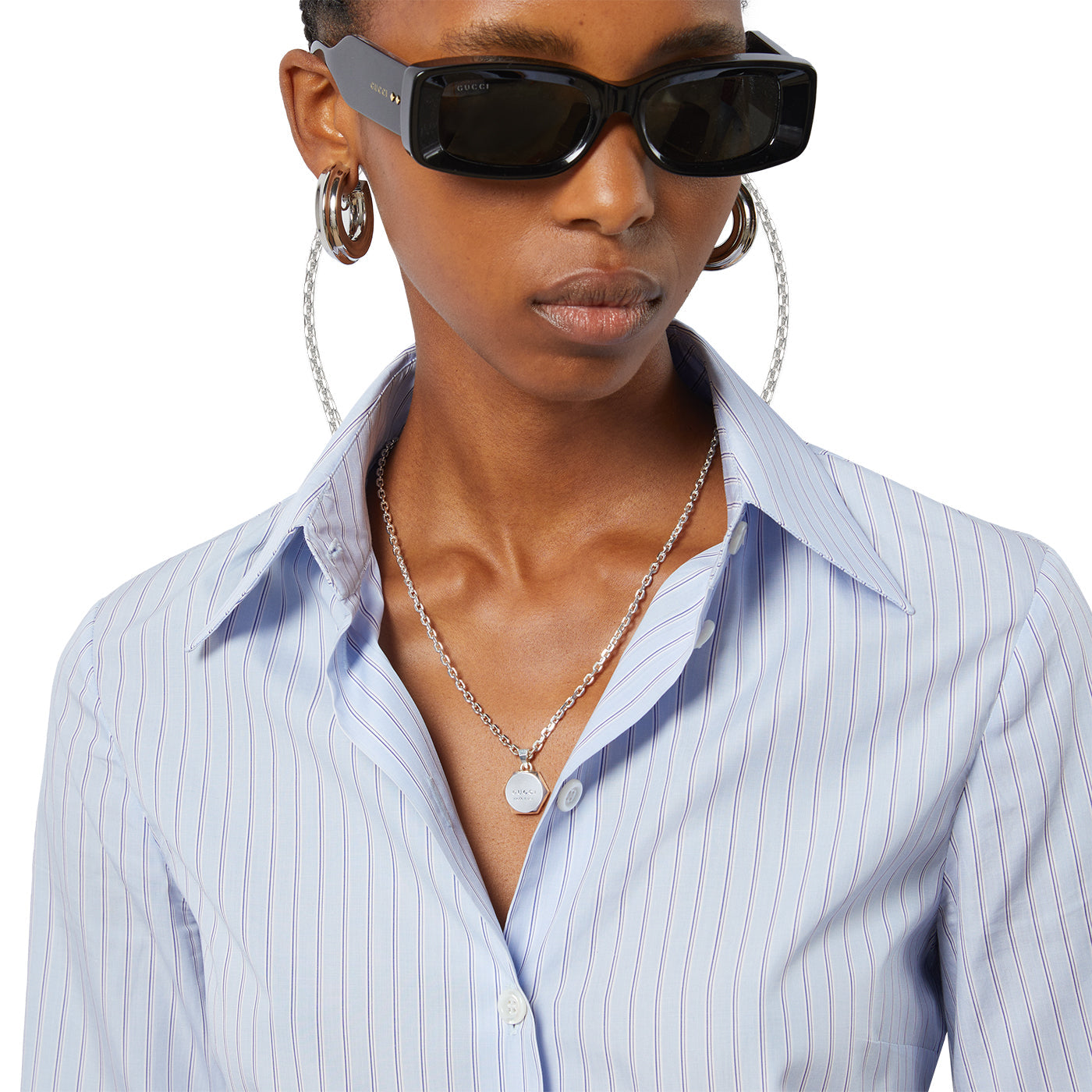 A person wears rectangular sunglasses, large hoop earrings, and a Gucci Trademark Sterling Silver Necklace with an adjustable length featuring a small pendant. They don a light blue, vertically striped collared shirt that complements their GUCCI Fine Jewellery style.