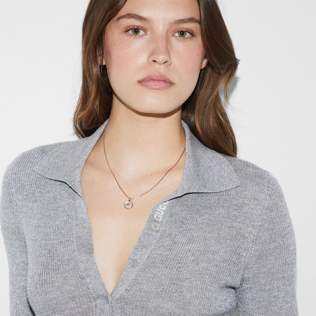 A woman with light brown hair wears a light gray, collared sweater and the GUCCI Fine Jewellery Interlocking G 18K Yellow Gold Diamond with Mother of Pearl Necklace Pendant, standing against a plain white background.