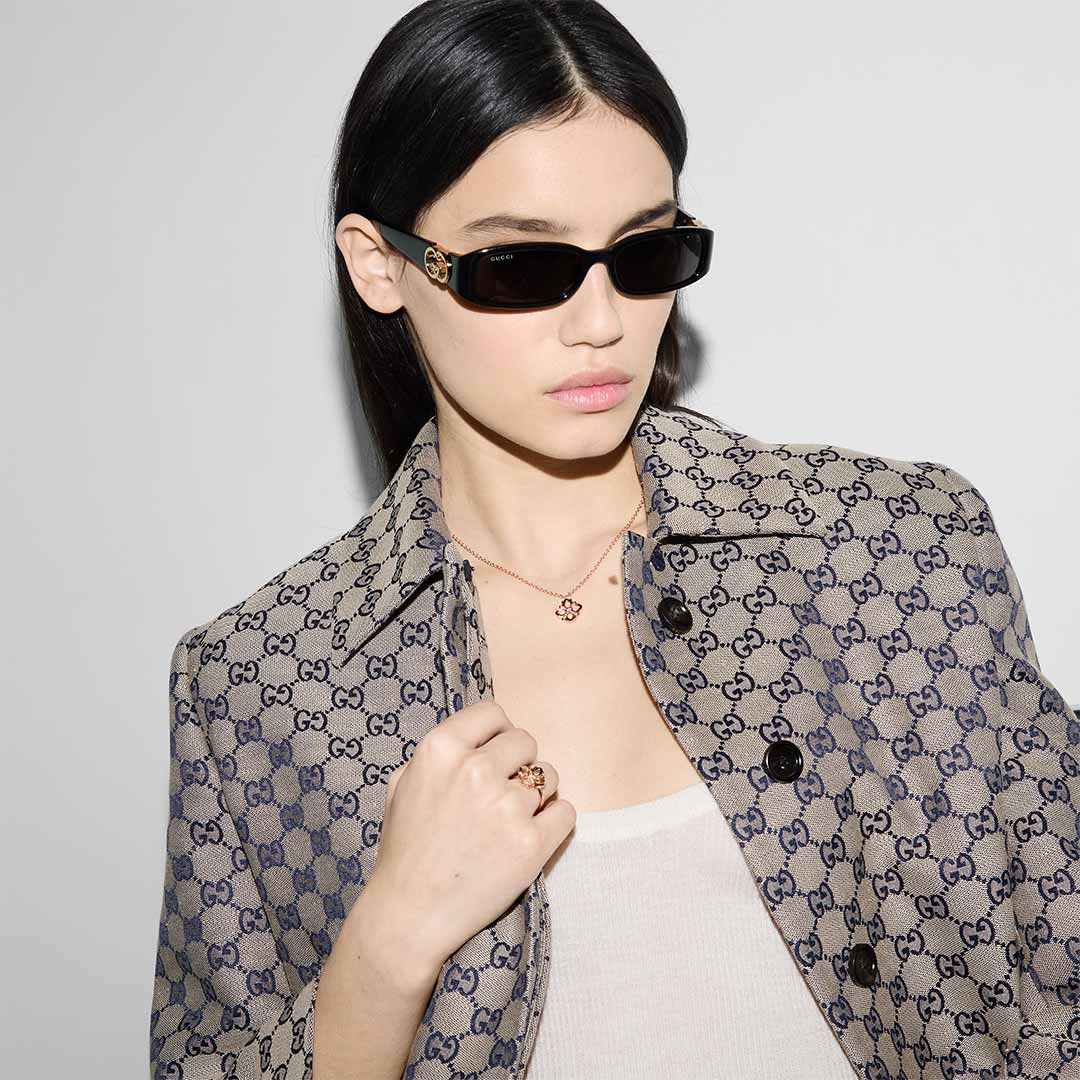 A person with long dark hair, wearing sleek dark sunglasses, a tan patterned jacket, and a white top enhances their look with the elegance of the Gucci Flora 18K Rose Gold Diamond Necklace Pendant by GUCCI Fine Jewellery. The sustainable jewelry stands out beautifully against a plain light background.