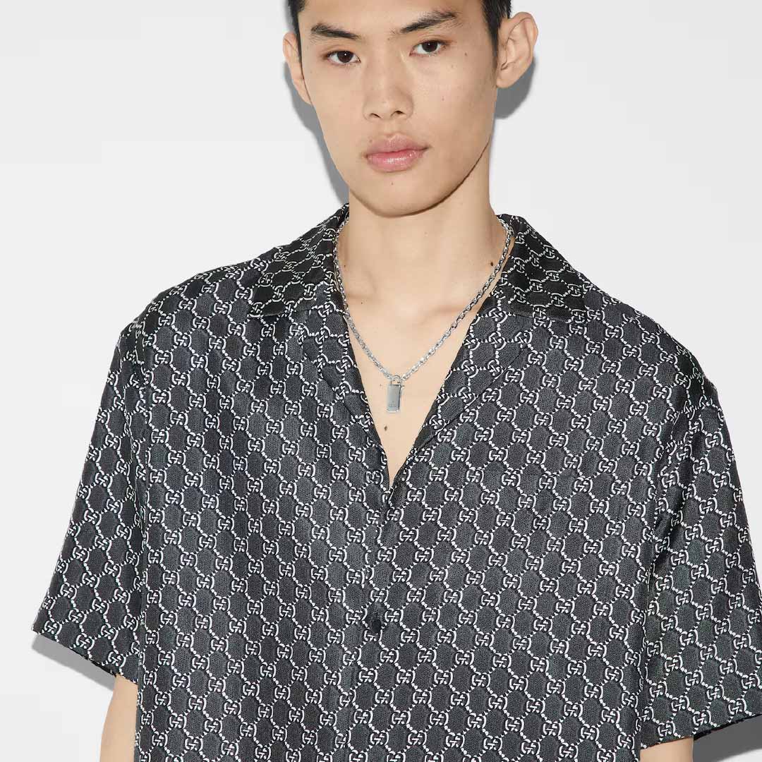 A person wearing a dark patterned shirt pairs it with the Gucci Trademark Sterling Silver Chain Necklace Pendant with Padlock from GUCCI Fine Jewellery, standing against a white background. The shirt's design, reminiscent of Gucci style, features a repeating pattern of interlocking shapes that captures the essence of Pre-Fall 2024 fashion.