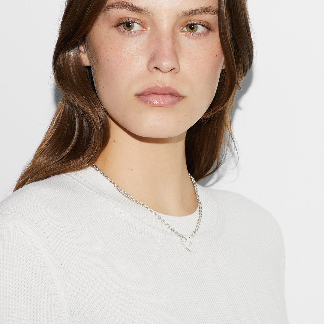 A person with long brown hair, wearing a simple white top and a GUCCI Fine Jewellery Trademark Sterling Silver Chain Necklace Pendant, stands against a plain white background. Their expression is neutral, maintaining a minimalistic look.