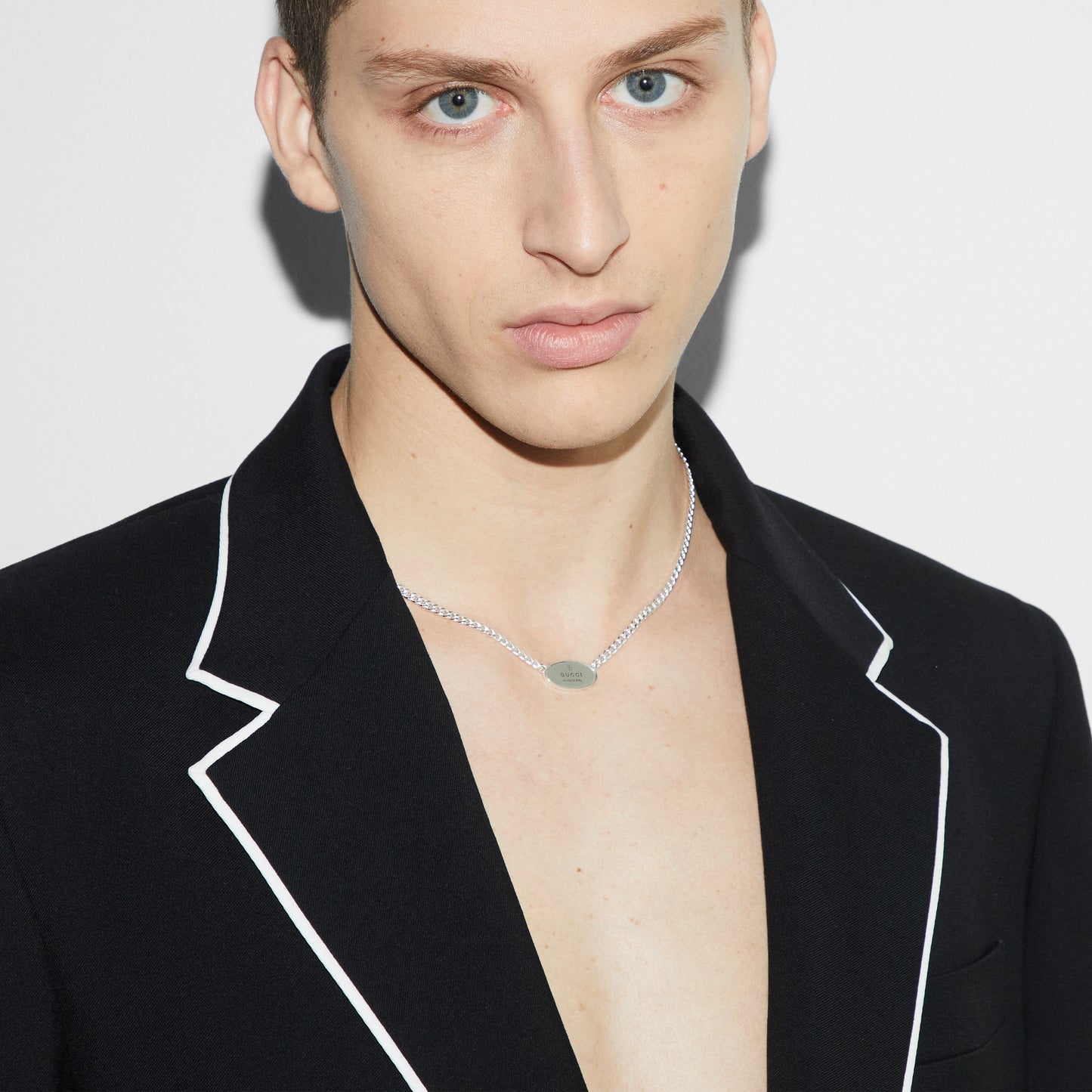 A person with short, light brown hair wears a black blazer with white trim and a Gucci Trademark Sterling Silver Tag Necklace Pendant from GUCCI Fine Jewellery. The plain white background highlights the person's neutral expression.