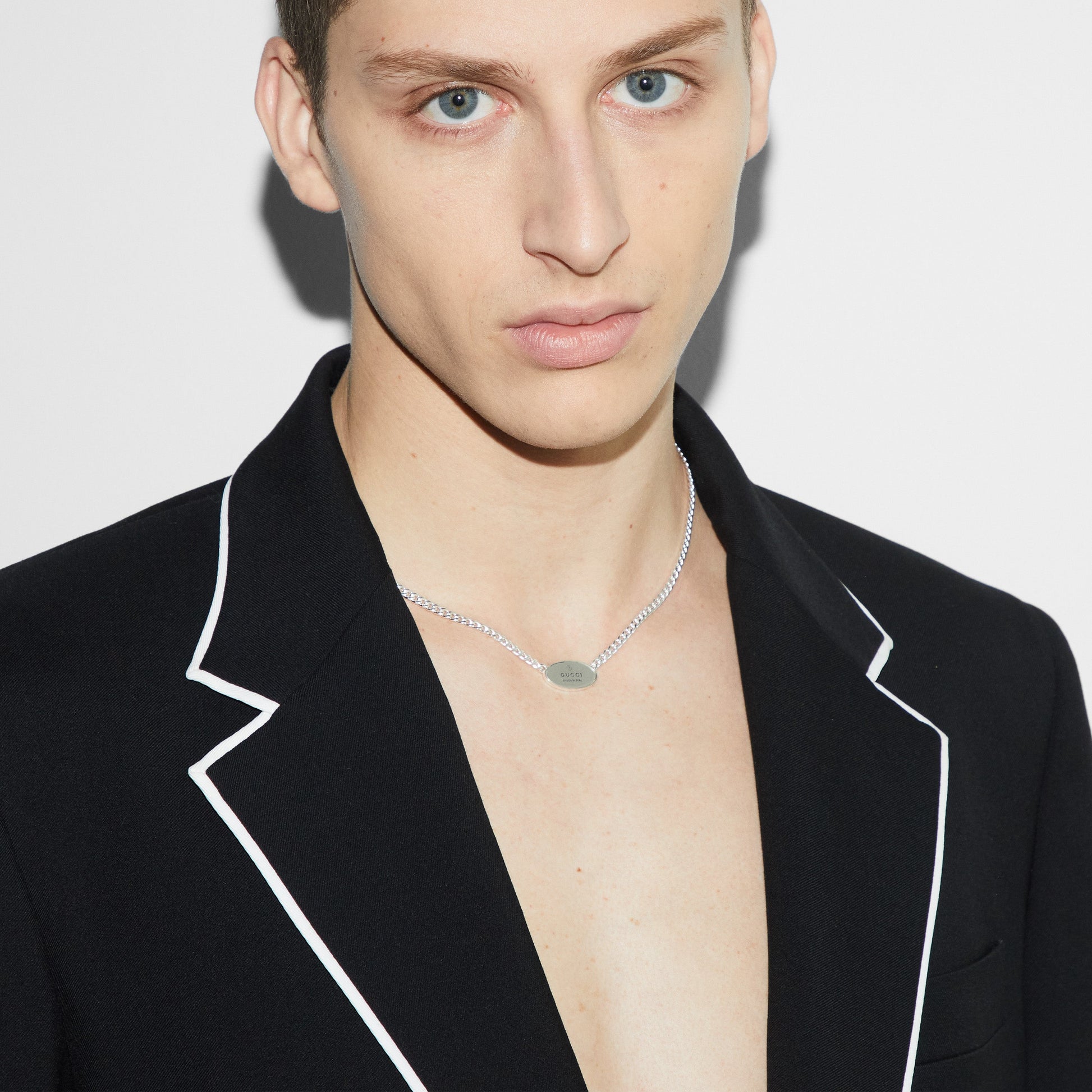 A person with short, light brown hair wears a black blazer with white trim and a Gucci Trademark Sterling Silver Tag Necklace Pendant from GUCCI Fine Jewellery. The plain white background highlights the person's neutral expression.