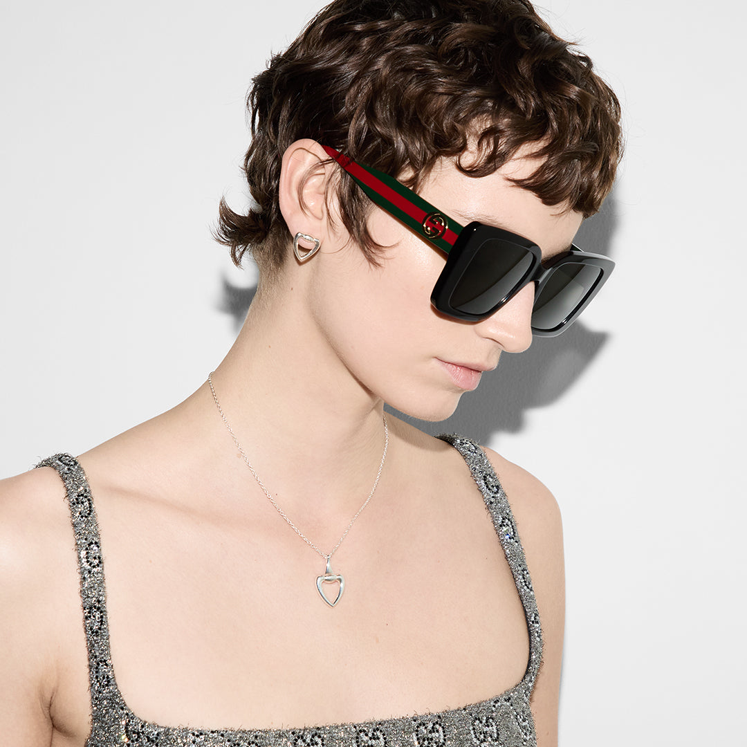 A person with short hair, large dark sunglasses, and a gray-patterned outfit models the Gucci Sterling Silver Heart Necklace Pendant by GUCCI Fine Jewellery against a plain white background.