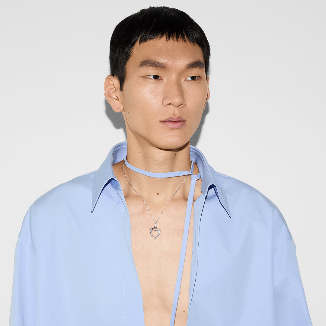A person wears a light blue shirt with an open collar, highlighting a Gucci Sterling Silver Heart Necklace Pendant from GUCCI Fine Jewellery. A thin blue ribbon is elegantly tied around the neck, complementing the subtle sparkle of the silver jewelry against a plain white background.