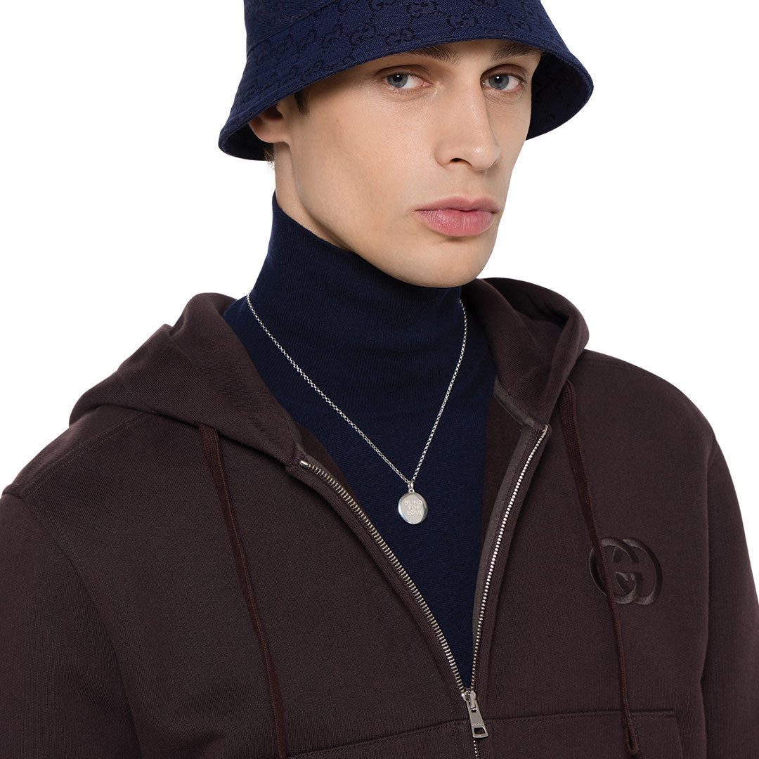 A person in a navy bucket hat, dark turtleneck, brown logo zip-up hoodie, and the GUCCI Fine Jewellery Blind For Love Sterling Silver Necklace Pendant, looking to the side against a white background.
