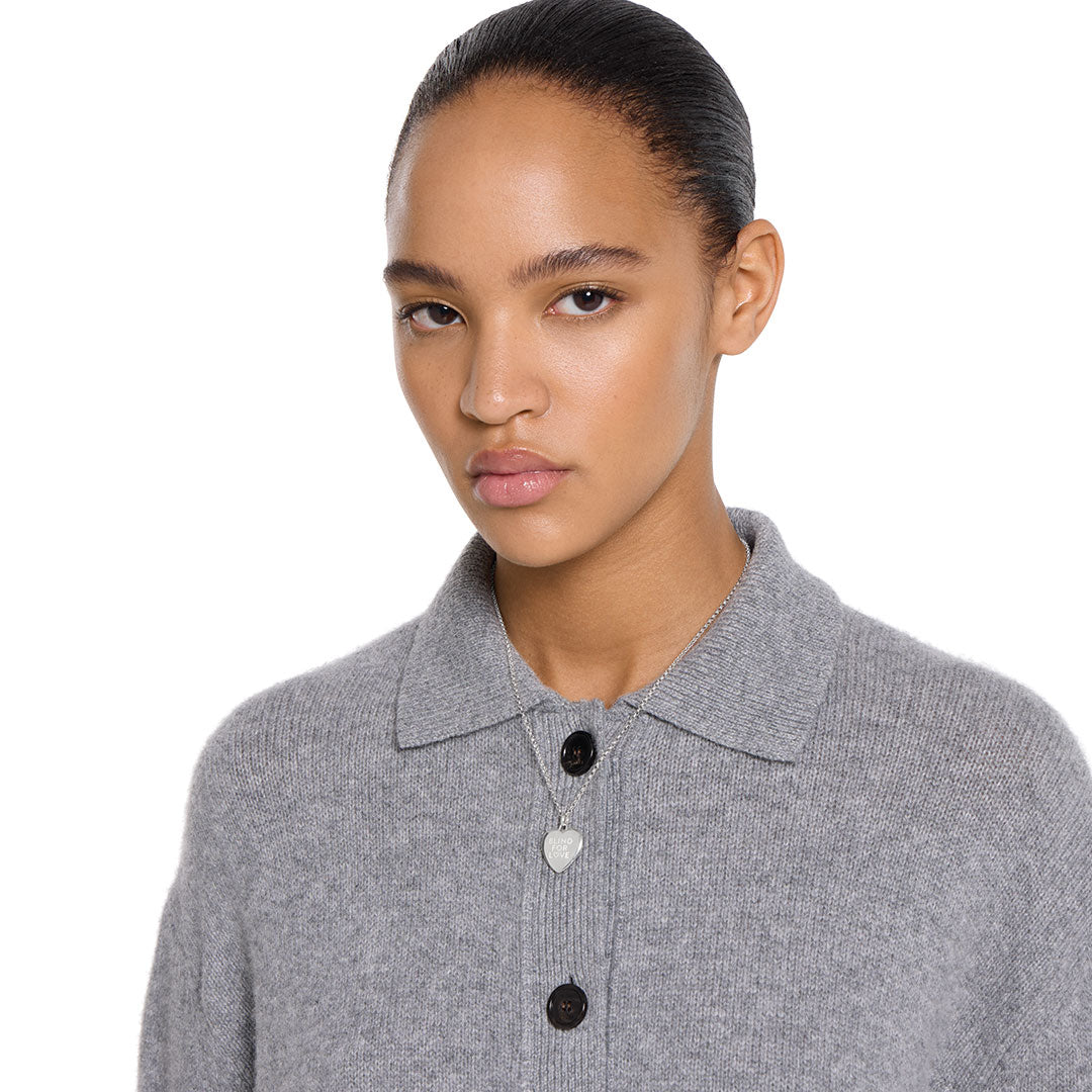 A woman with smooth, pulled-back hair wears a gray button-up sweater and the GUCCI Fine Jewellery Blind For Love Sterling Silver Necklace Pendant, standing against a plain white background.