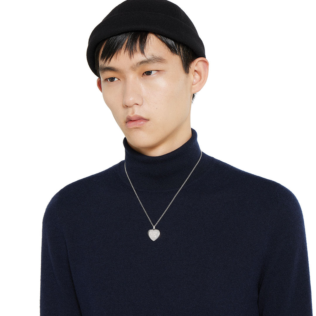 A person with straight black hair, in a black beanie and navy turtleneck, wears a GUCCI Fine Jewellery "Blind For Love" sterling silver necklace pendant, looking slightly to the side against a plain white background.