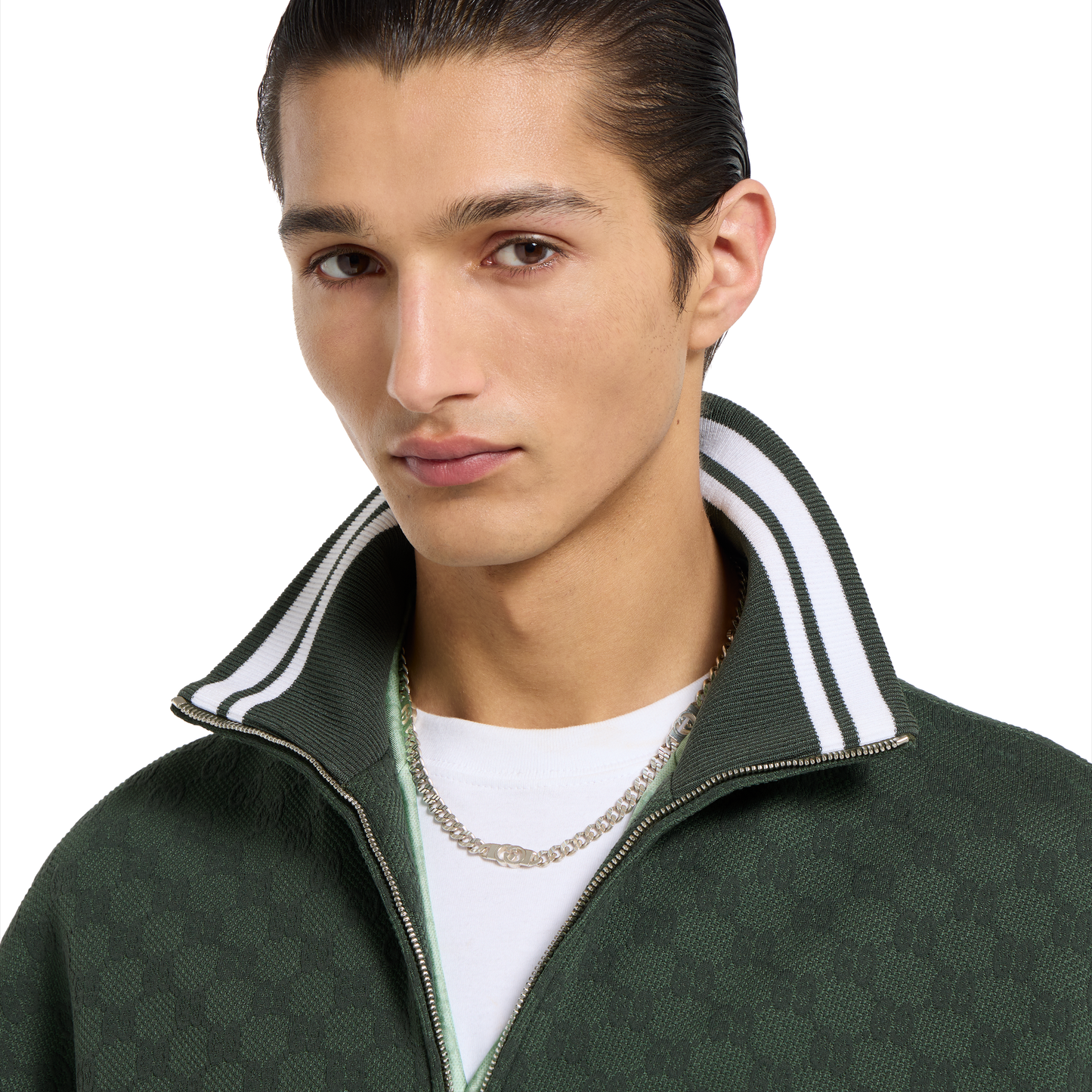 A young man with slicked-back dark hair wears a dark green zip-up jacket with a white and green striped collar, a white shirt, and the Gucci Interlocking Sterling Silver Chain Necklace from GUCCI Fine Jewellery, looking at the camera with a neutral expression.