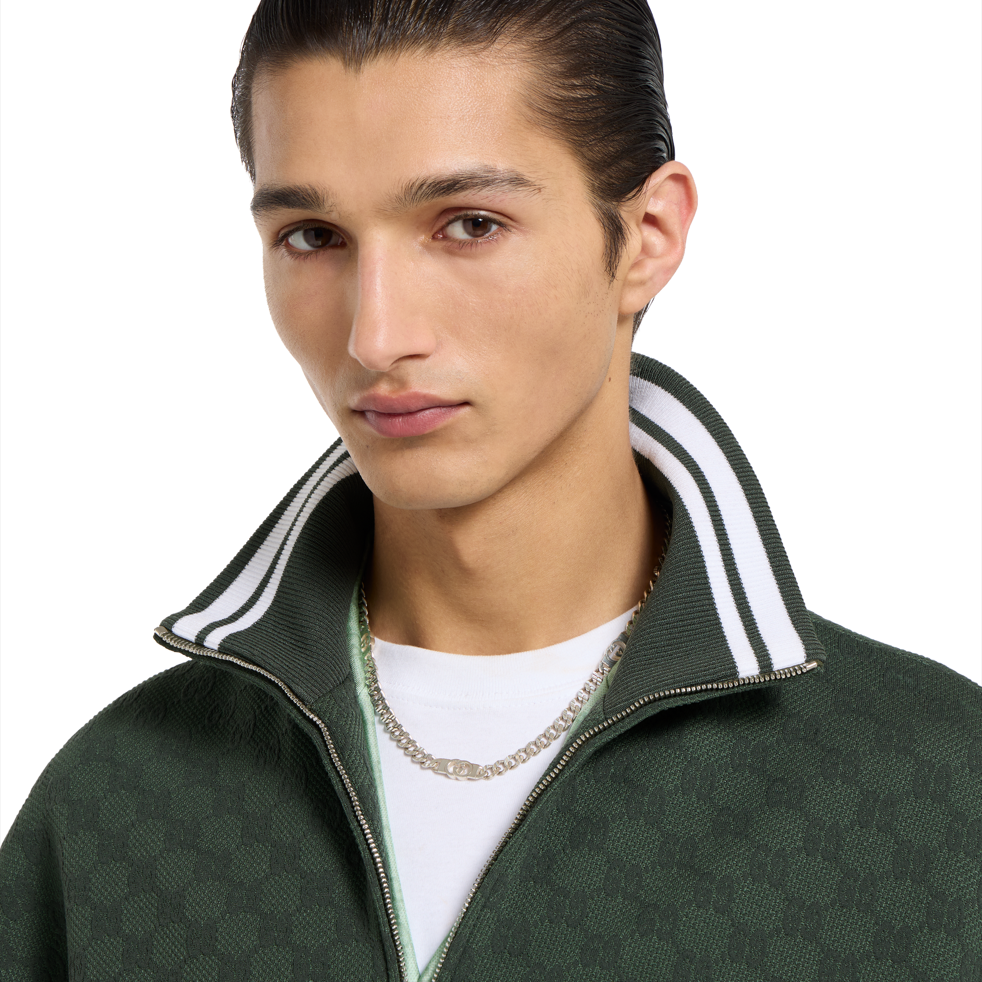A young man with slicked-back dark hair wears a dark green zip-up jacket with a white and green striped collar, a white shirt, and the Gucci Interlocking Sterling Silver Chain Necklace from GUCCI Fine Jewellery, looking at the camera with a neutral expression.
