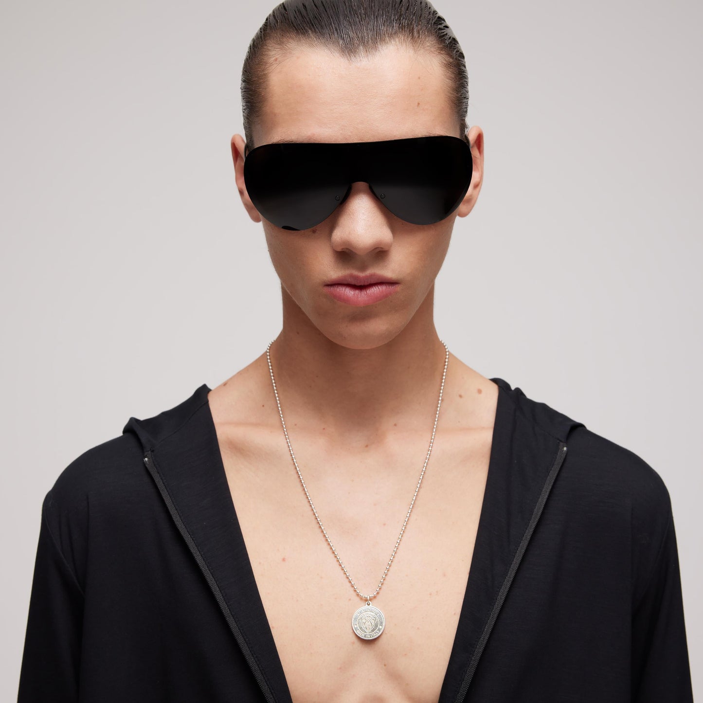 A person with slicked-back hair wears large black futuristic sunglasses, a black open shirt, and a GUCCI Fine Jewellery Gucci Crest Sterling Silver Necklace Pendant, posing against a plain light background.
