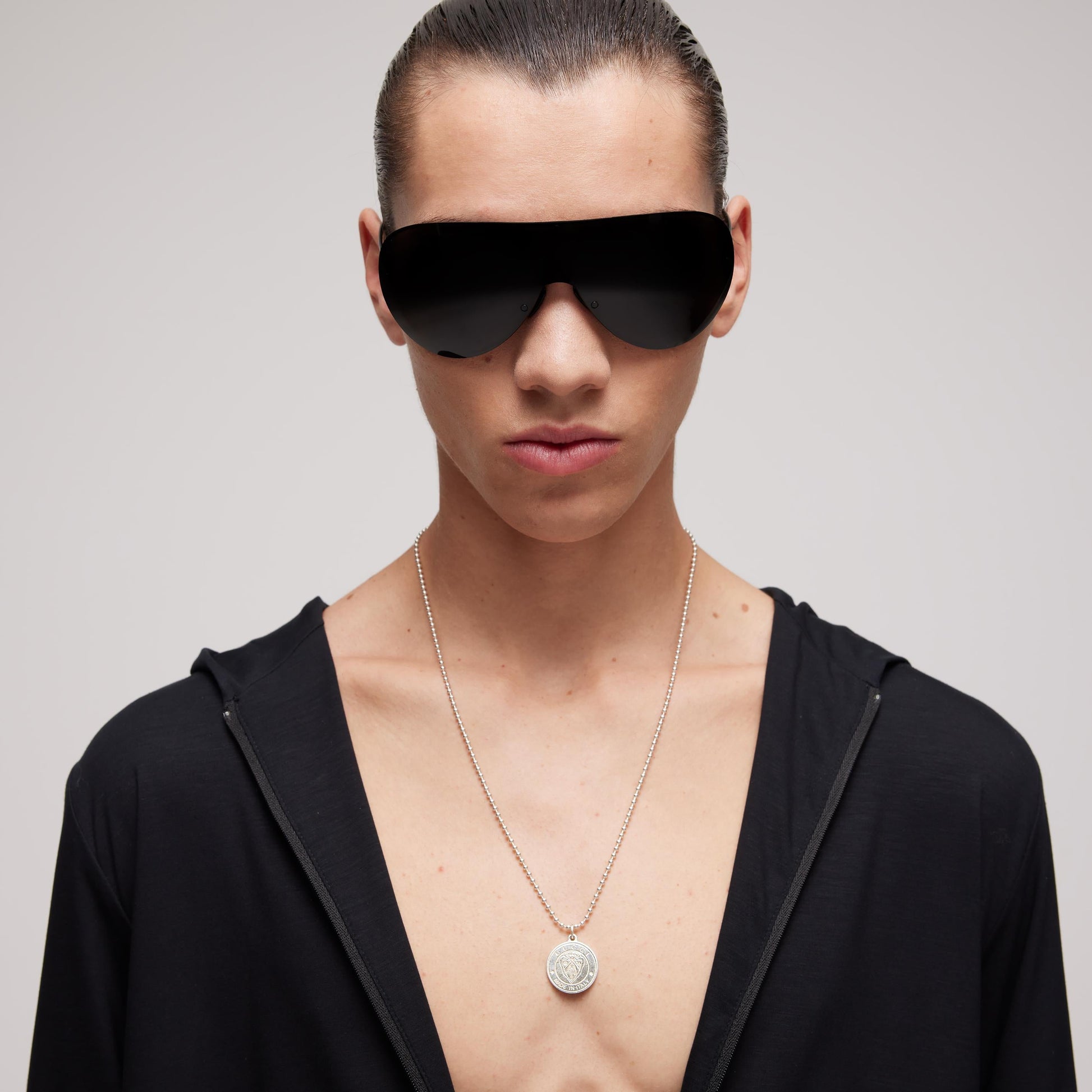 A person with slicked-back hair wears large black futuristic sunglasses, a black open shirt, and a GUCCI Fine Jewellery Gucci Crest Sterling Silver Necklace Pendant, posing against a plain light background.
