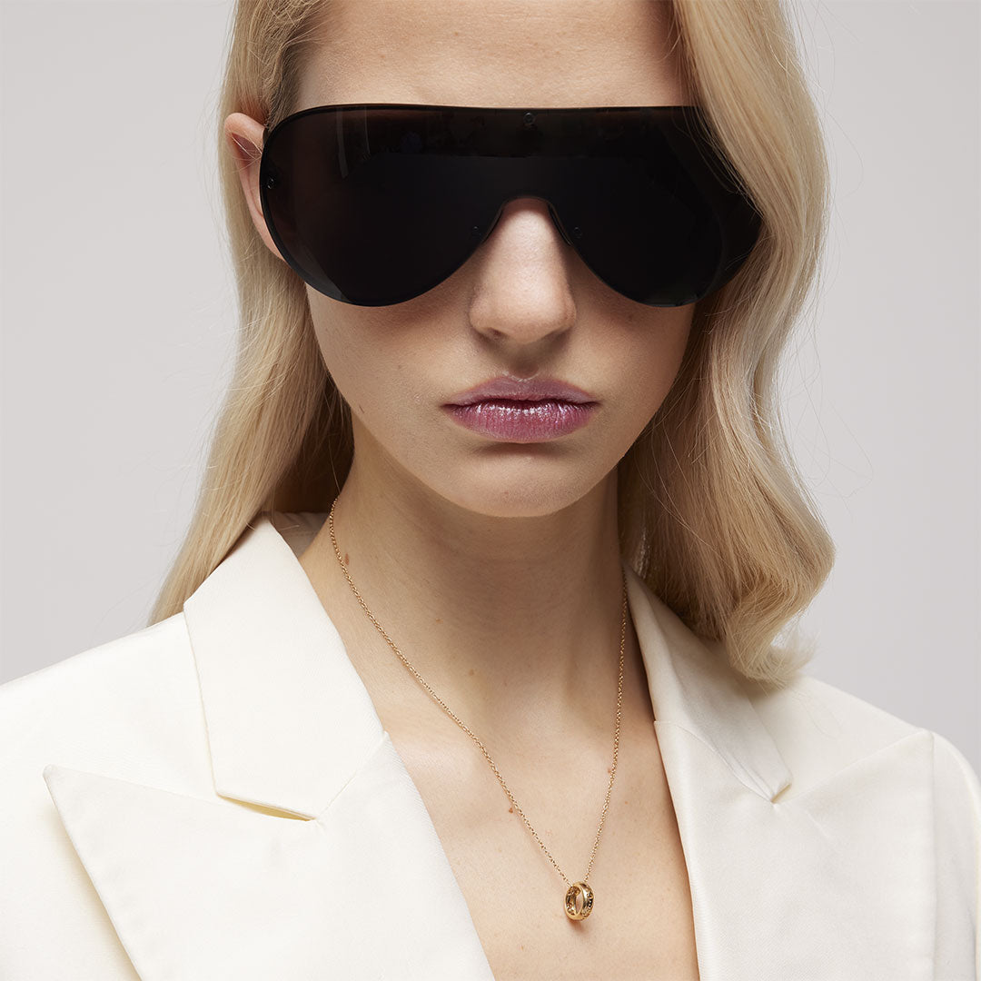 A woman with long blonde hair wears large black sunglasses, a white blazer, and the GUCCI Fine Jewellery Gucci Icon 18K Yellow Gold Necklace Pendant featuring the signature GG cut-out motif, standing against a plain light background.