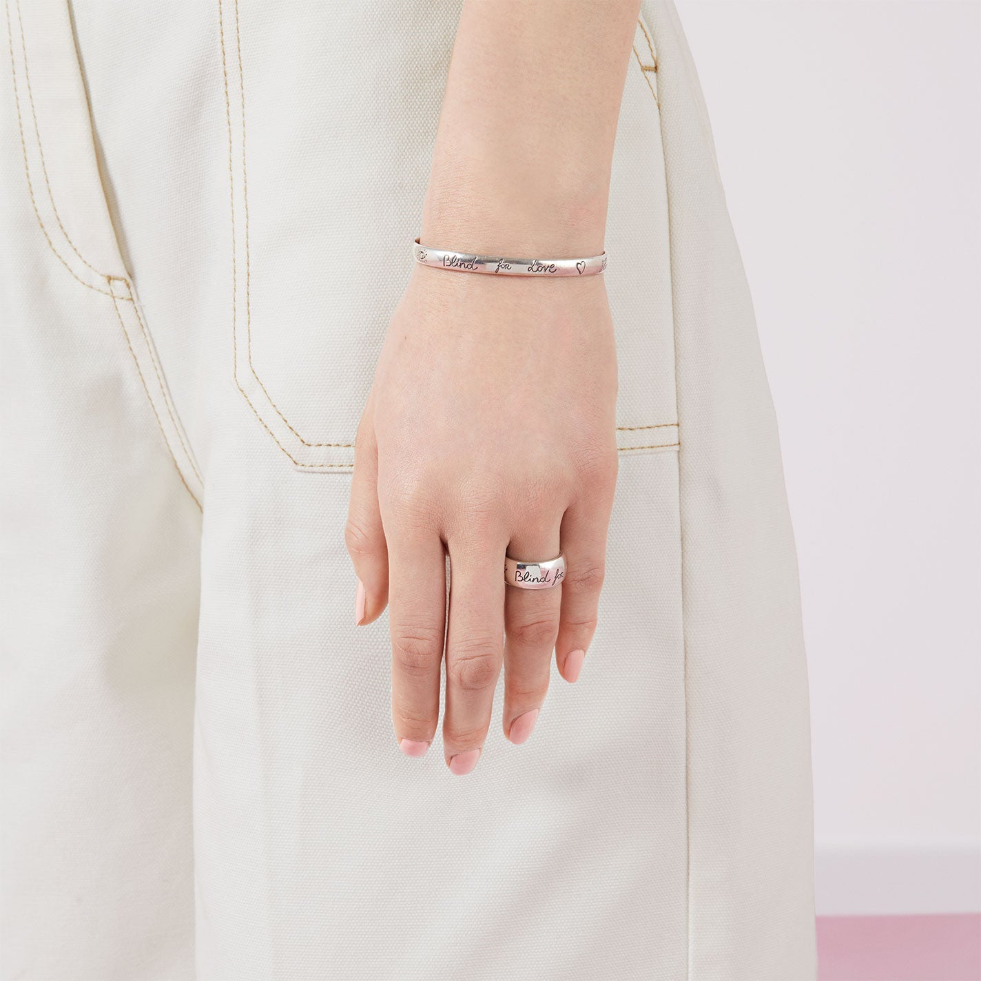 A person adorned with a Gucci Blind For Love Sterling Silver Ring by GUCCI Fine Jewellery is also wearing a bracelet from the same collection. They are dressed in white pants, with their arm resting alongside their body against a soft pink and white wall.