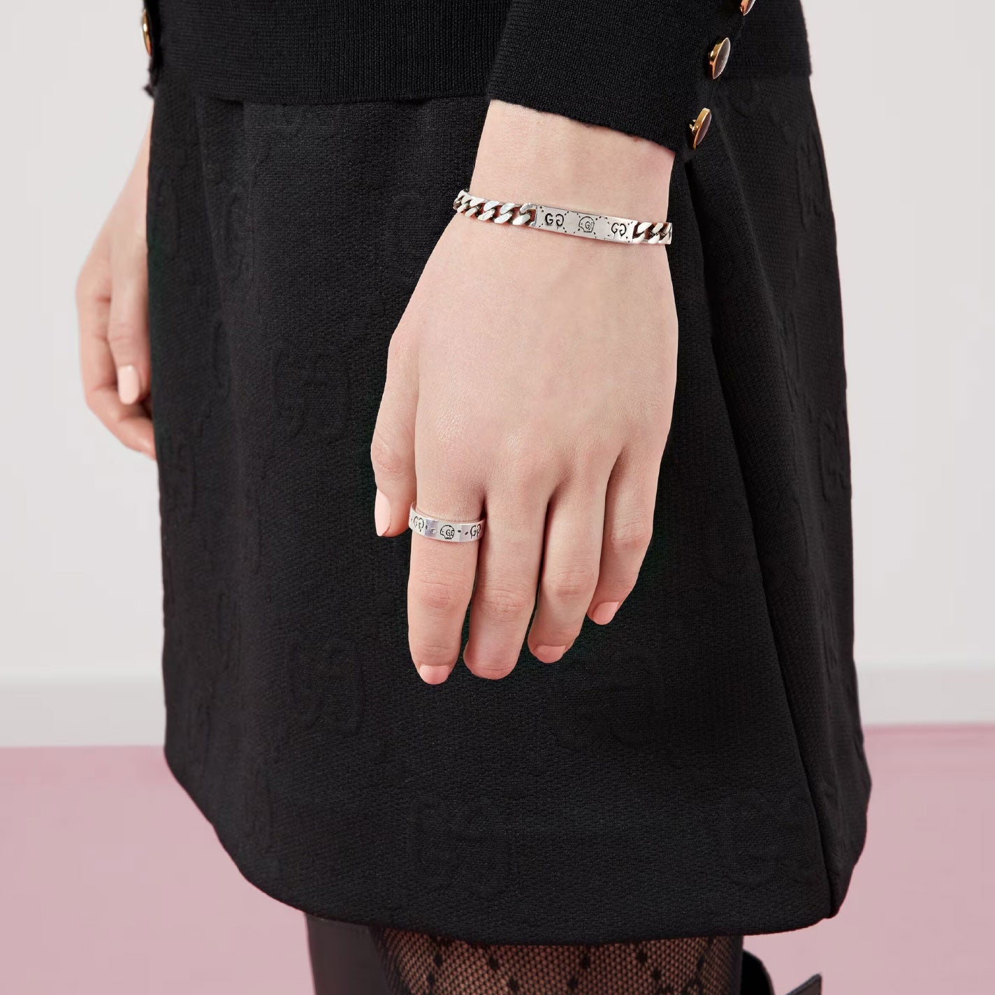 A person dressed in a black dress showcases their hand, adorned with a Gucci Ghost Sterling Silver Ring and a matching chain-link bracelet, set against a backdrop of a white wall and pink floor.