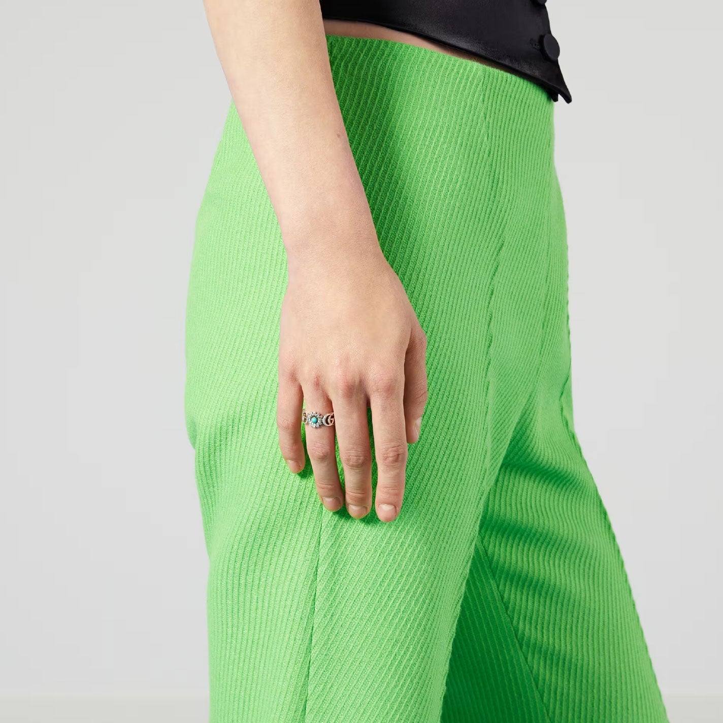 A person wearing bright green ribbed pants and a black top stands against a neutral background. Their hand, adorned with the Gucci Double G Sterling Silver Ring with Mother of Pearl, Blue Topaz, and Turquoise Resin by GUCCI Fine Jewellery, gleams as it catches the light.