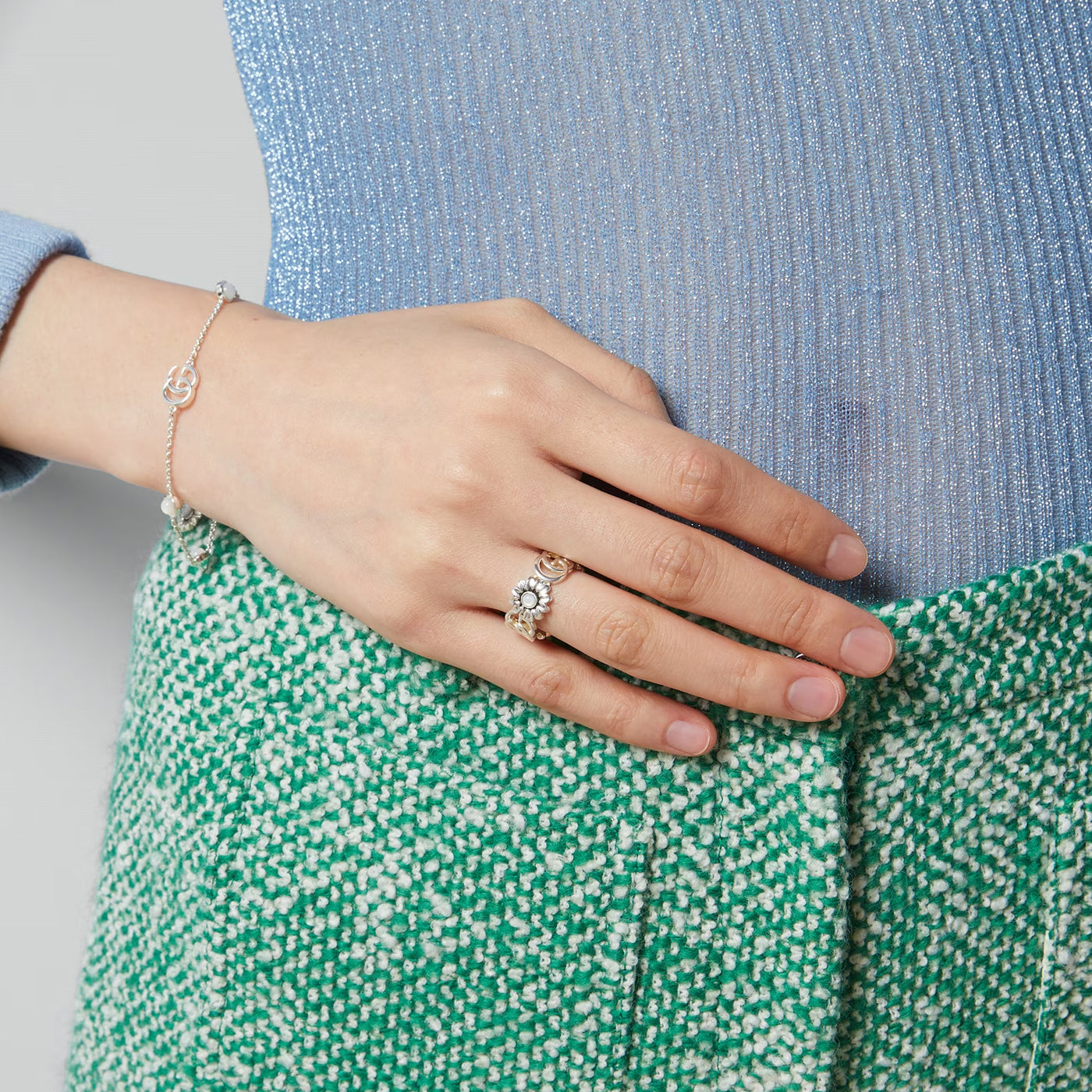 A hand adorned with the Gucci GG Marmont Sterling Silver Mother of Pearl Ring by GUCCI Fine Jewellery rests on a hip, complemented by a matching bracelet. The outfit features a sparkly blue top and a textured green and white skirt, showcasing the elegance of fashion jewellery.