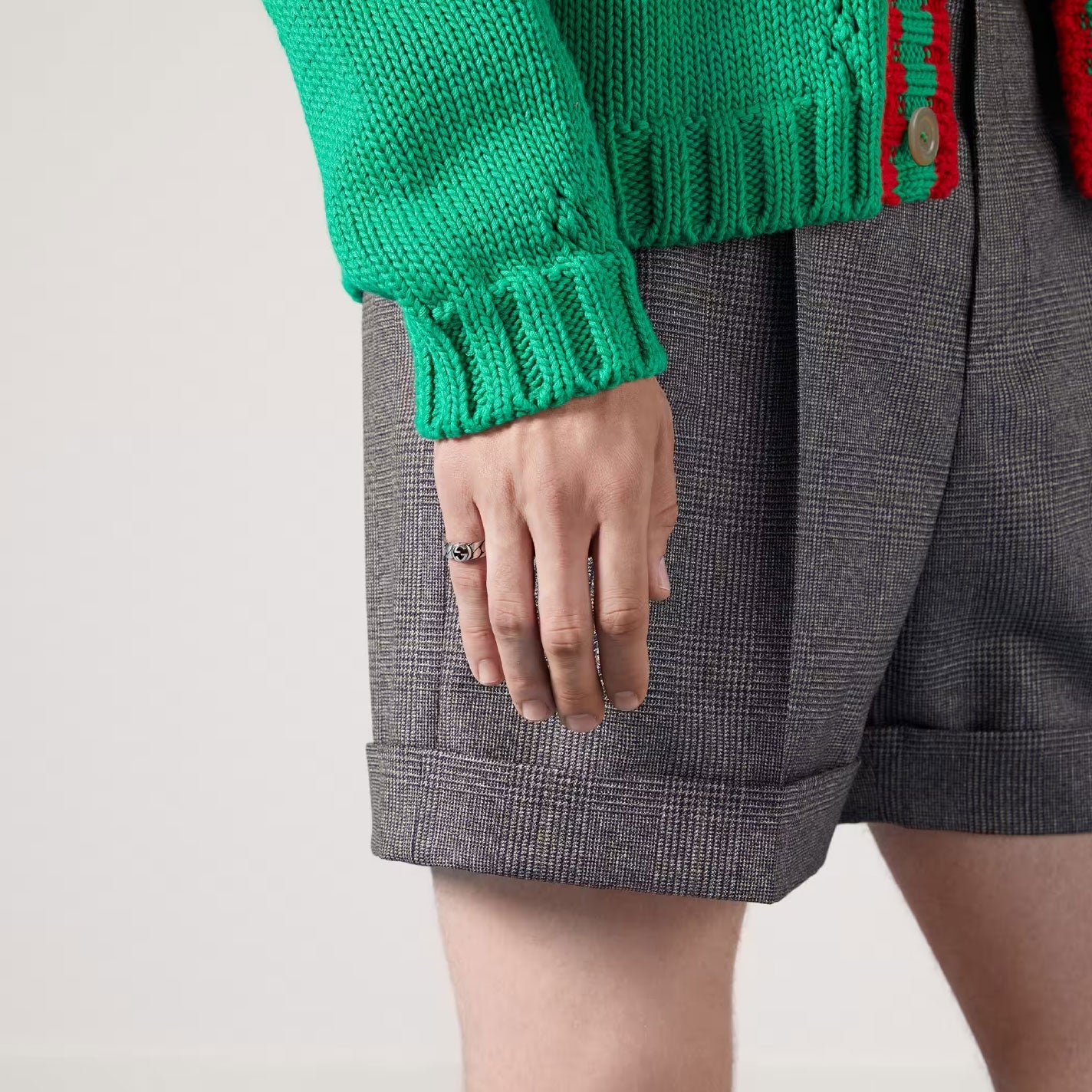 A person wears a vibrant green knit sweater paired with gray plaid shorts. Their hand, adorned with a Gucci Interlocking G Sterling Silver Ring by GUCCI Fine Jewellery, rests at their side. Drawing from vintage inspirations, the look effortlessly stands out against the plain light backdrop.