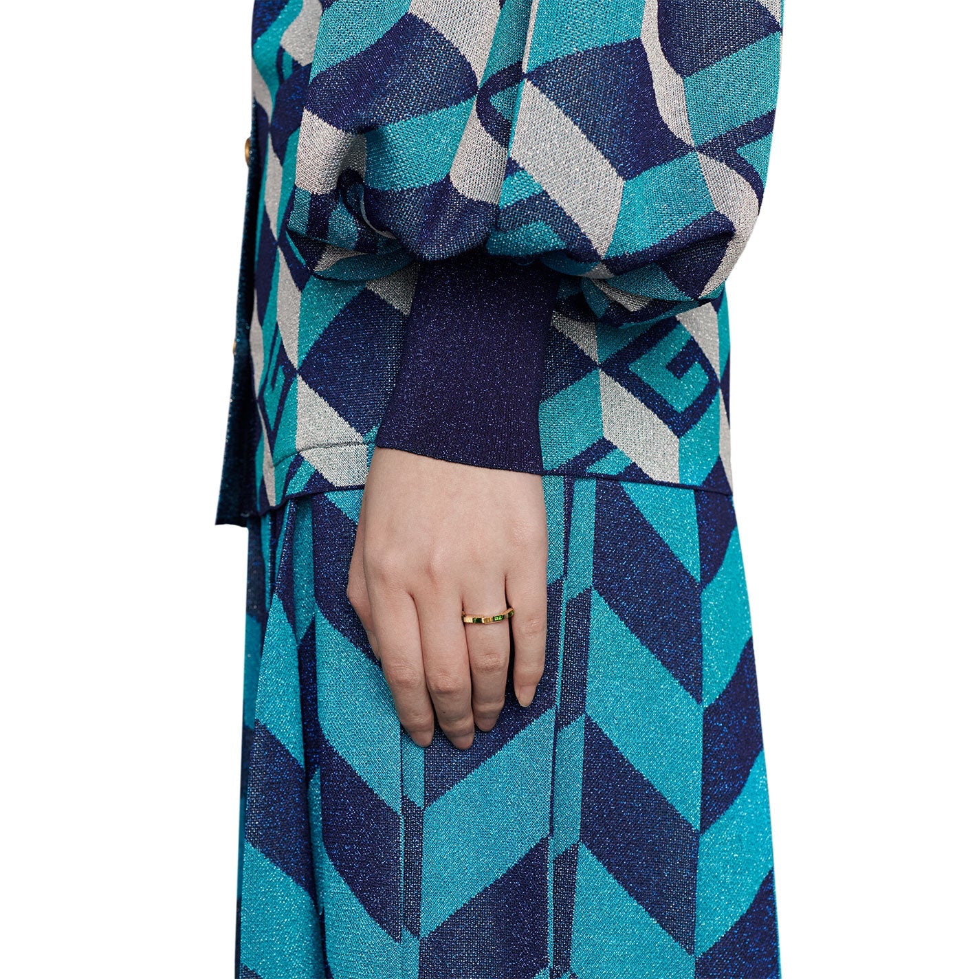 A person is dressed in a vibrant, geometric-patterned outfit in shades of blue and teal. On their hand is a Gucci Link to Love Tourmaline 18K Yellow Gold Ring by GUCCI Fine Jewellery. Their ensemble includes a long-sleeved top and a matching skirt.