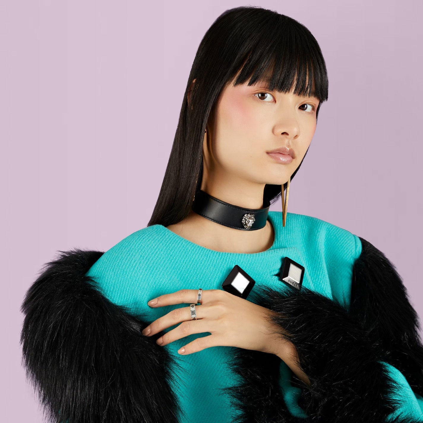 A person with long black hair and bangs, dressed in a turquoise outfit with black fur trim and a black choker necklace, poses against a light purple background. They have a serious expression and are accessorized with long earrings and the Gucci GG And Bee Engraved Thin Sterling Silver Ring from GUCCI Fine Jewellery.