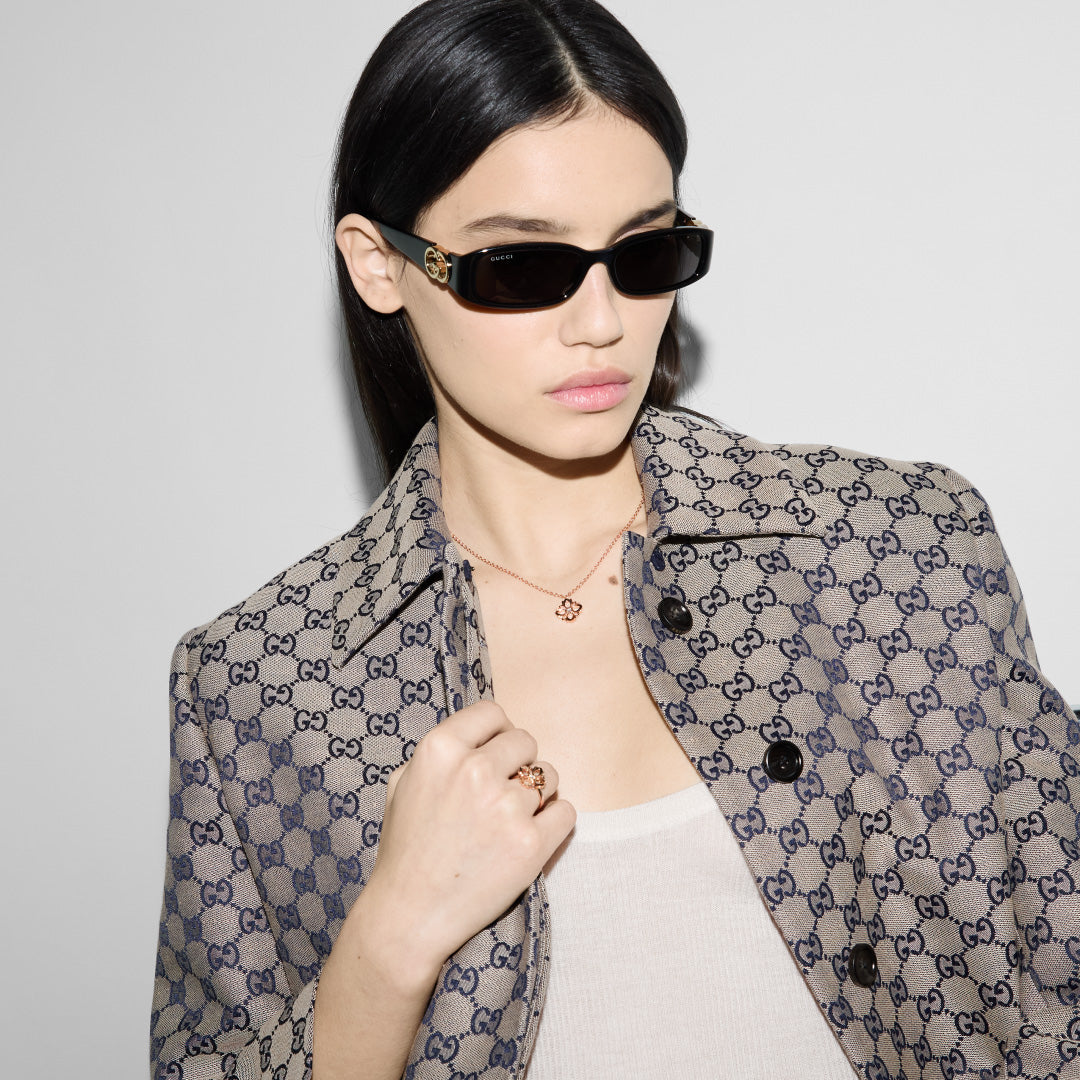 A woman wearing dark sunglasses and a Gucci-patterned jacket poses against a plain background. She accessorizes with the Gucci Flora 18K Rose Gold Diamond Fashion Ring from GUCCI Fine Jewellery. Her expression is neutral, highlighting the chic elegance of her ensemble.
