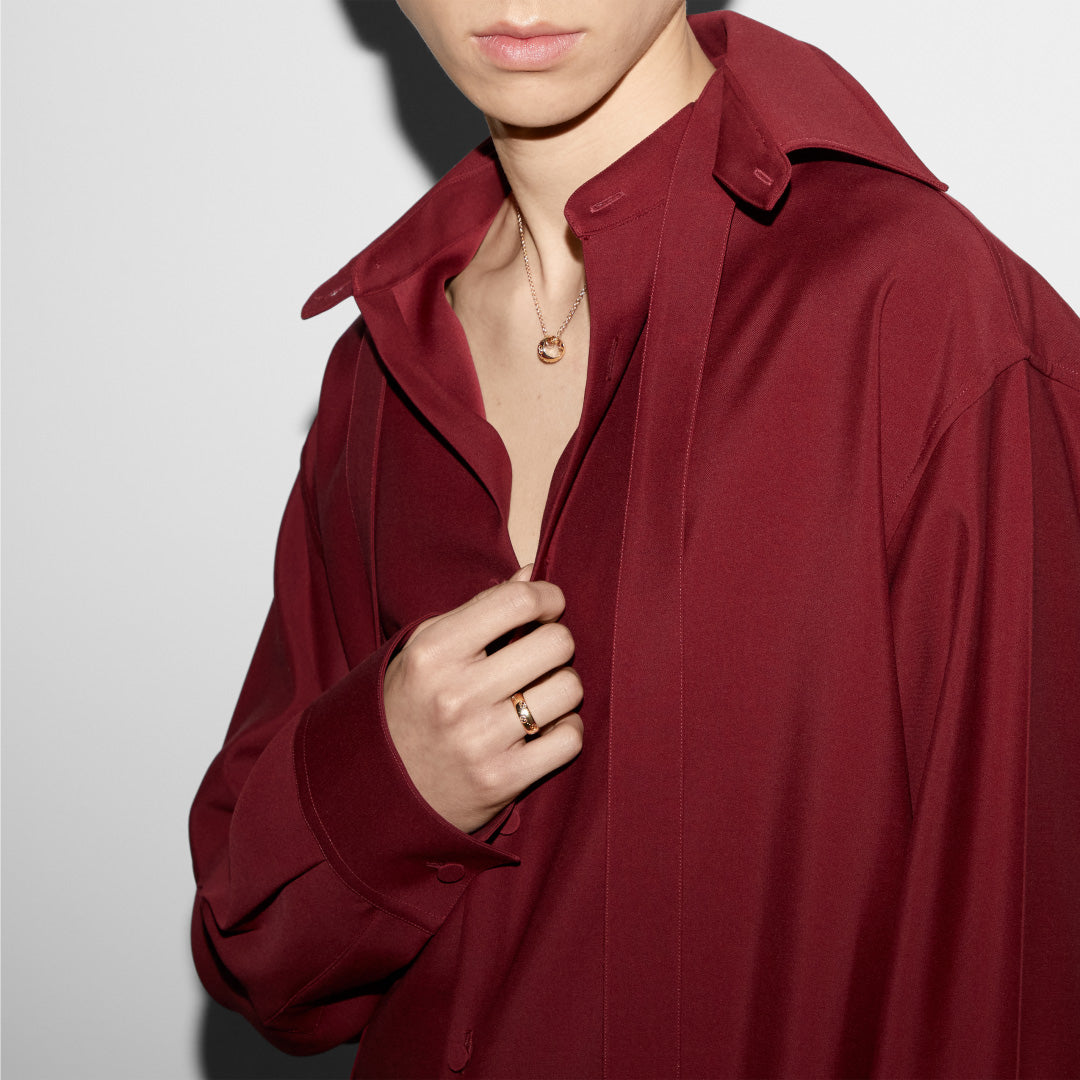 A person is wearing a deep red shirt with the collar open, showcasing a Gucci jewelry necklace, along with a Gucci Icon GG 18K Yellow Gold GG Thin Ring on their hand. The emphasis is on the clothing and exquisite accessories, set against a plain white background.