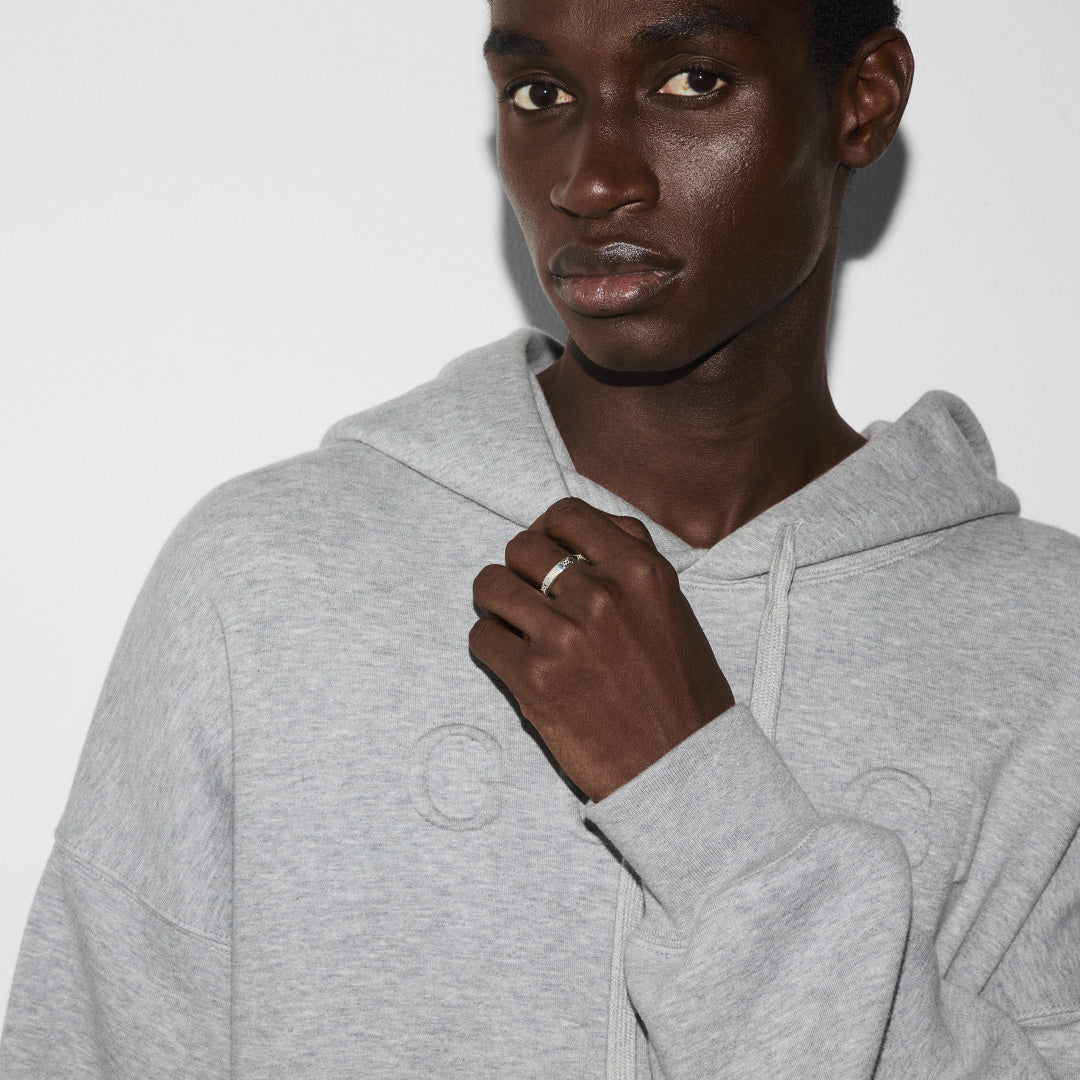 Someone wearing a light gray hoodie stands against a plain white background, subtly showcasing a Gucci Signature GG Sterling Silver Thin Ring. They grip the hoodie’s drawstrings with one hand, revealing a touch of sterling silver from the GUCCI Fine Jewellery collection. Their neutral expression is accentuated by soft shadows in the lighting.