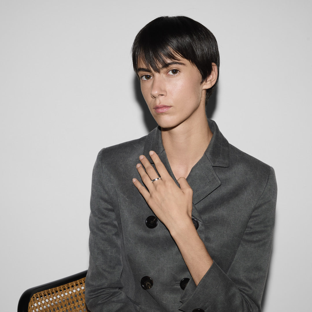 A person with short dark hair sits against a light background, wearing a gray double-breasted blazer. Their hand, adorned with a Gucci Signature GG Sterling Silver Thin Ring from GUCCI Fine Jewellery, gently rests on their chest. They are seated on a chair with a woven backrest.