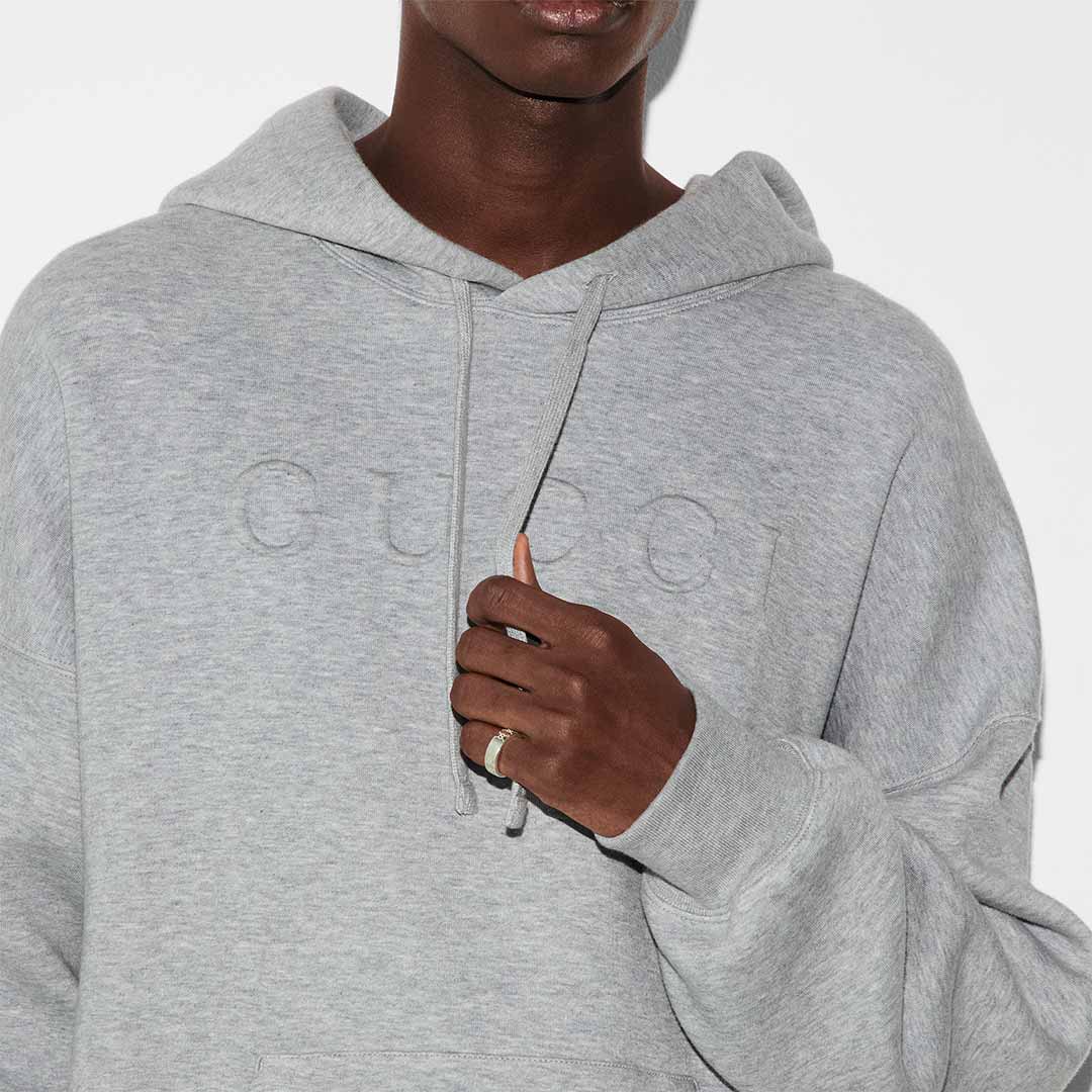 A person dressed in a light gray hoodie featuring a subtle embossed design on the front, captured from the neck down. They hold the hoodie’s drawstring with one hand and wear a Gucci Signature GG Sterling Silver Wide Ring by GUCCI Fine Jewellery.