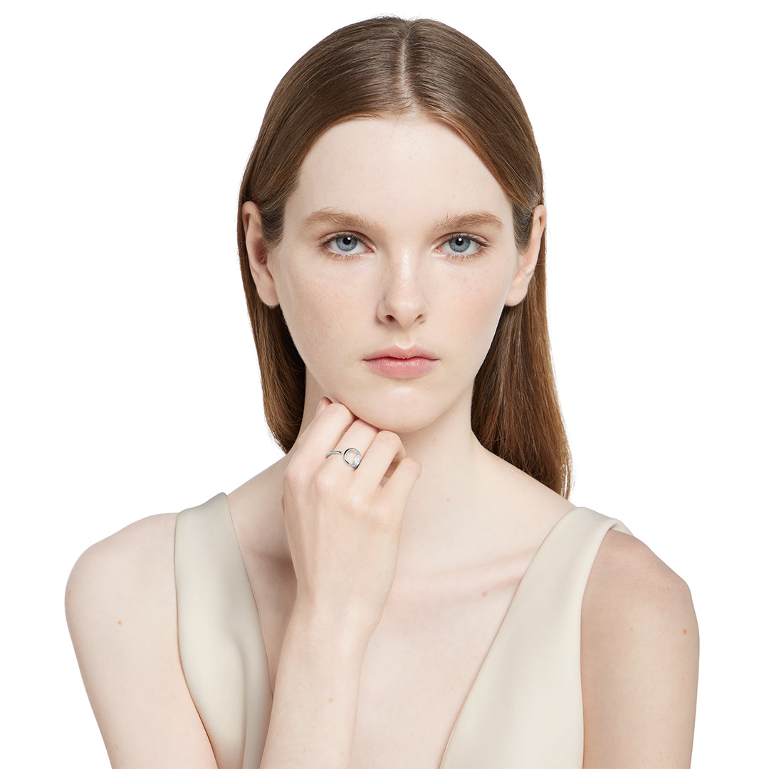 A young woman with fair skin and straight brown hair poses against a white background, wearing a sleeveless beige top and showcasing the GUCCI Fine Jewellery Horsebit 18K White Gold Diamond Ring on her left hand as she gently touches her chin.