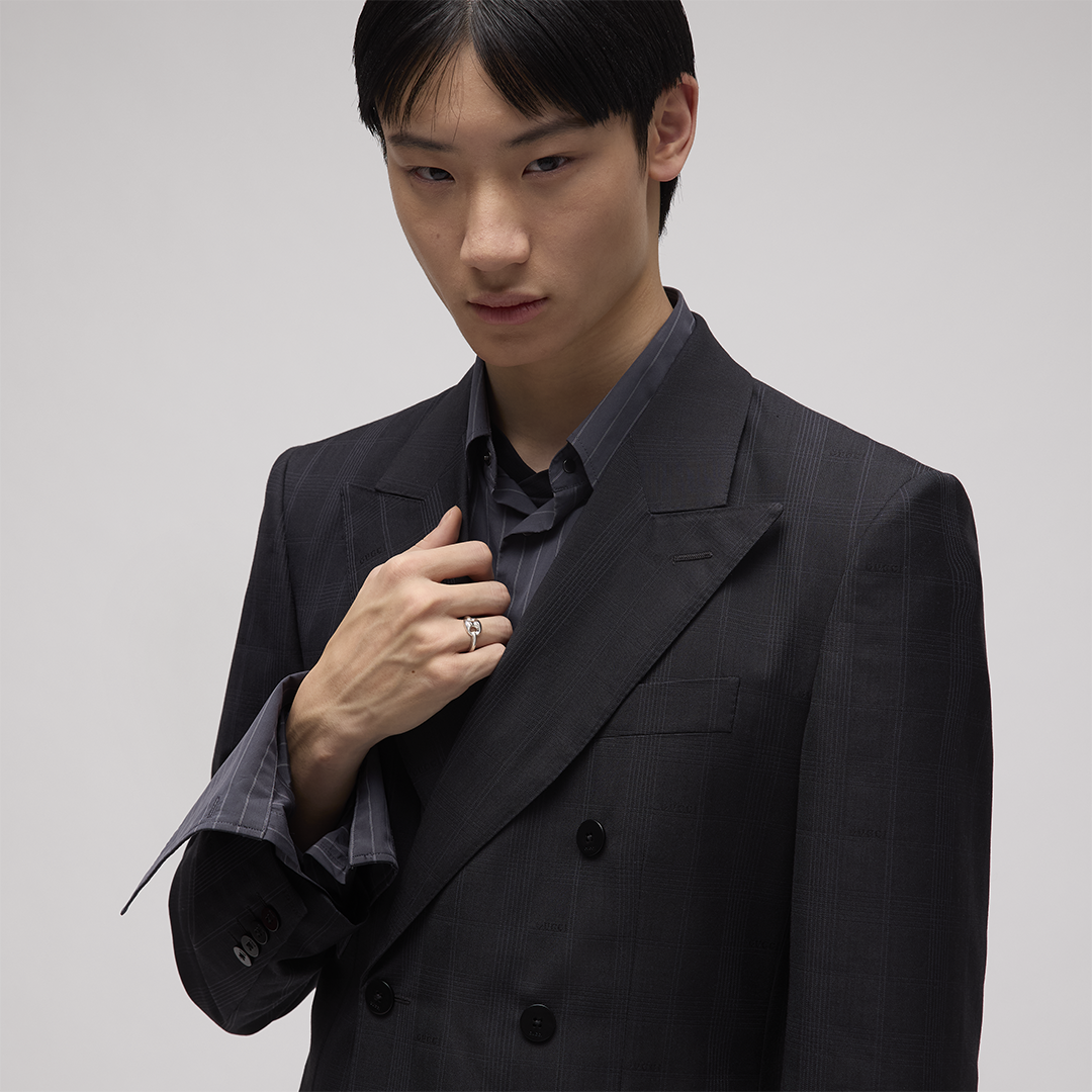A person in a dark suit jacket holds their lapel to showcase a Gucci Marina Sterling Silver Chain Ring on their finger.
