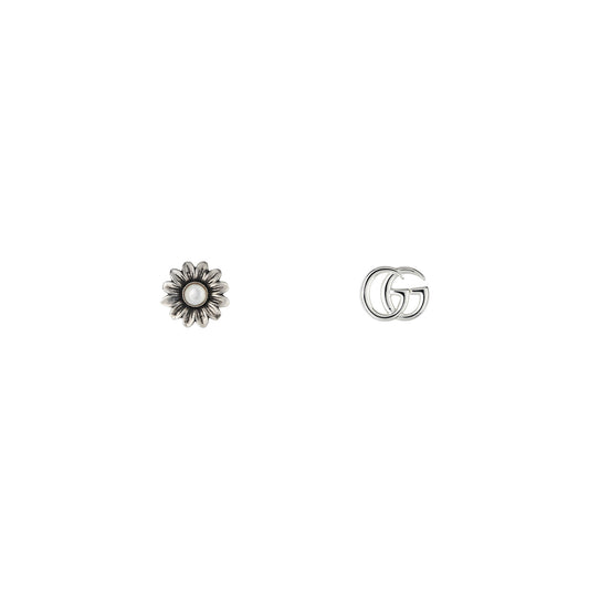 A pair of stud earrings from GUCCI Fine Jewellery is displayed on a white background: one earring is intricately designed as a flower with a mother of pearl center, while the other showcases the iconic Double G logo. Both are crafted from sterling silver, radiating an elegant Gucci charm.