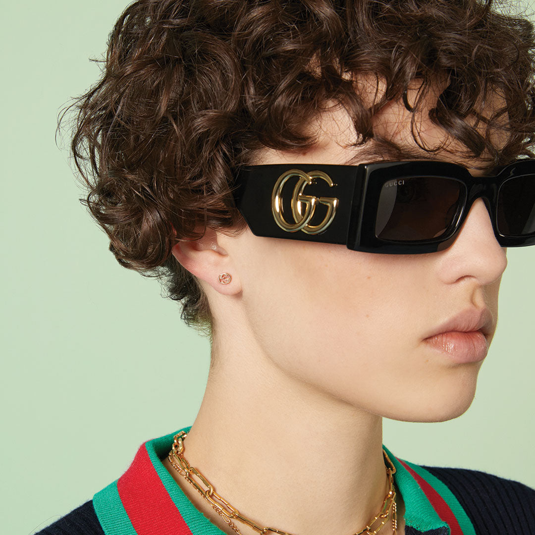 A person with short curly brown hair wears large black sunglasses, a green and red collared shirt, a navy sweater, and Gucci Interlocking G 18K Rose Gold Stud Earrings by GUCCI Fine Jewellery against a light green background.