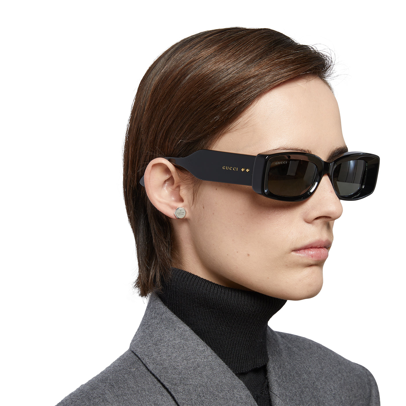 A person with short hair is wearing large, black rectangular sunglasses and a gray blazer over a black turtleneck, accentuated by Gucci Trademark Sterling Silver Stud Earrings from GUCCI Fine Jewellery.