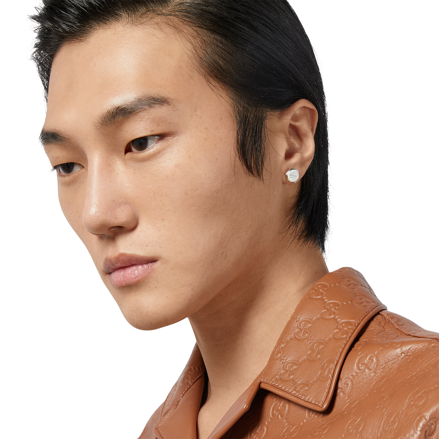 A person with short black hair is wearing a light brown leather jacket and a small, geometric Gucci Trademark Sterling Silver Stud Earring by GUCCI Fine Jewellery. The plain white background enhances the stylish simplicity of the ensemble.