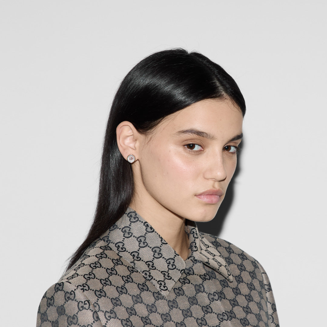 A person with long dark hair and Gucci Interlocking 18K Rose Gold Diamond Stud Earrings from GUCCI Fine Jewellery is gazing intently. They are wearing a patterned Gucci shirt featuring the Interlocking G, standing against a plain white background.