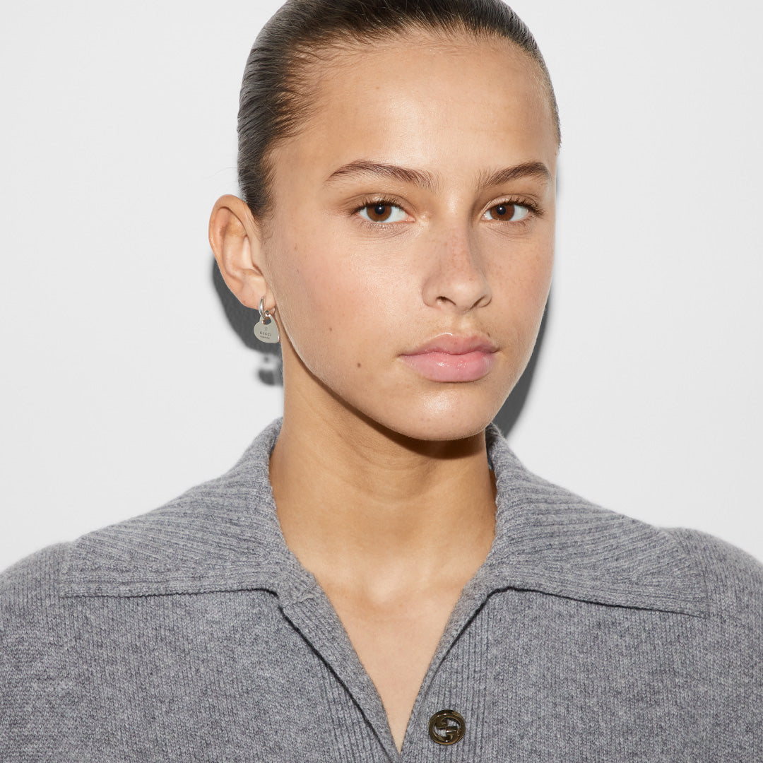 A person with a neutral expression is wearing a gray collared sweater and a small Gucci Trademark Sterling Silver Hoop Earring. Their hair is pulled back, and they stand against a plain white background, accessorized with stylish pieces from GUCCI Fine Jewellery.