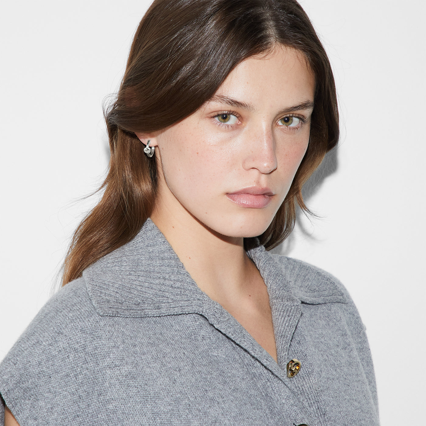 A person with long brown hair is wearing a gray collared sweater and the Gucci Interlocking G Sterling Silver Hoop Earrings by GUCCI Fine Jewellery. They are looking directly at the camera against a plain white background.