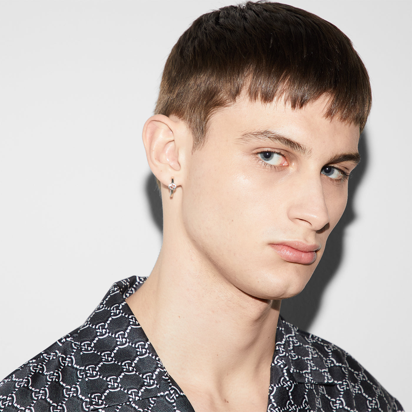 A young person with short brown hair and light skin, wearing a patterned black and white shirt, is looking to the side. They have the Gucci Interlocking G Sterling Silver Hoop Earrings from GUCCI Fine Jewellery adorning their left ear, set against a plain white background.