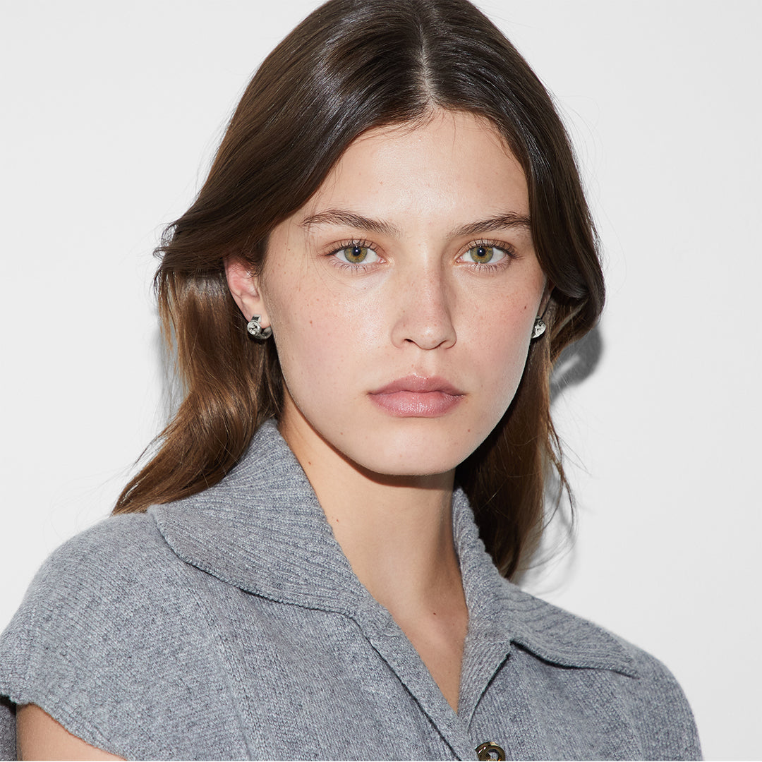 A woman with long brown hair and light eyes wears a gray collared top and GUCCI Fine Jewellery Gucci Interlocking G Sterling Silver Wide Hoop Earrings, looking directly at the camera against a plain white background.