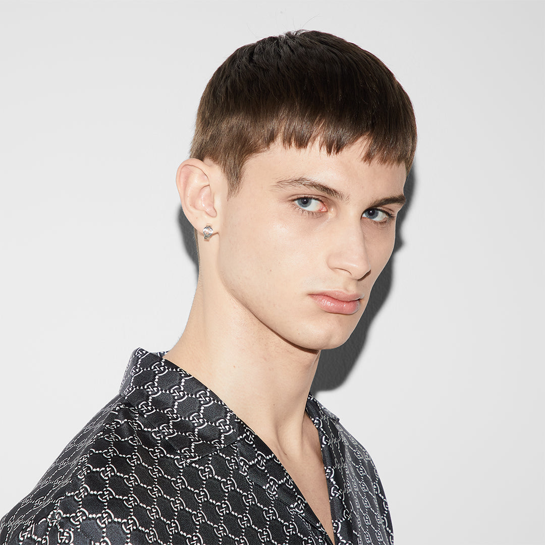 A young man with short brown hair and blue eyes, wearing a black patterned shirt and a GUCCI Fine Jewellery Interlocking G Sterling Silver Wide Hoop Earring in his left ear, stands against a plain white background.