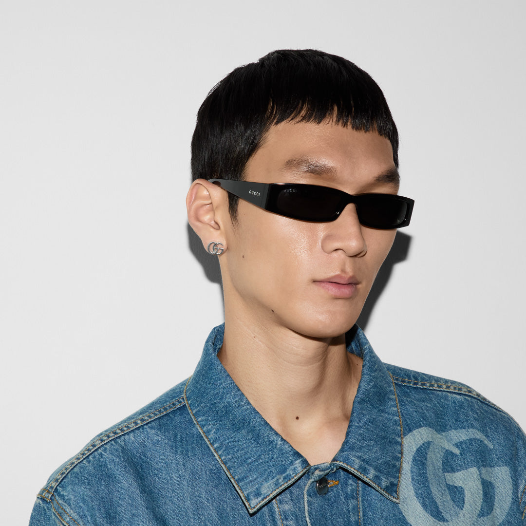 A person wearing rectangular black sunglasses and a denim jacket stands in front of a plain white background. The jacket features a large, stylized "G" reminiscent of the GG Marmont style, while the person sports jewelry from GUCCI Fine Jewellery with a small Gucci GG Mormont Sterling Silver Stud Earring visible on one ear.