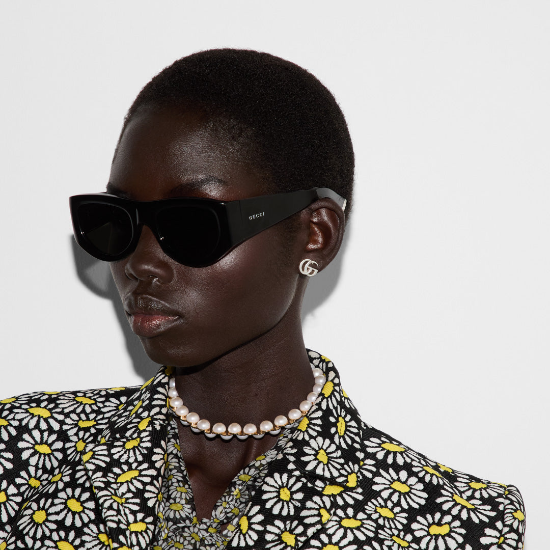 A person adorned in black sunglasses, a pearl necklace, and Gucci GG Mormont Sterling Silver Stud Earrings by GUCCI Fine Jewellery, elegantly donning a floral-patterned outfit with yellow, white, and black daisies against a plain white background.