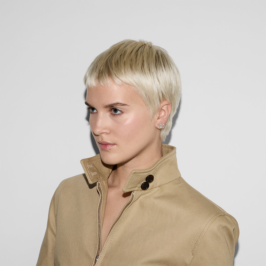 A person with short platinum blonde hair and light skin wears a beige collared jacket and GUCCI Fine Jewellery Interlocking Sterling Silver Stud Earrings, looking slightly to the side against a plain white background.