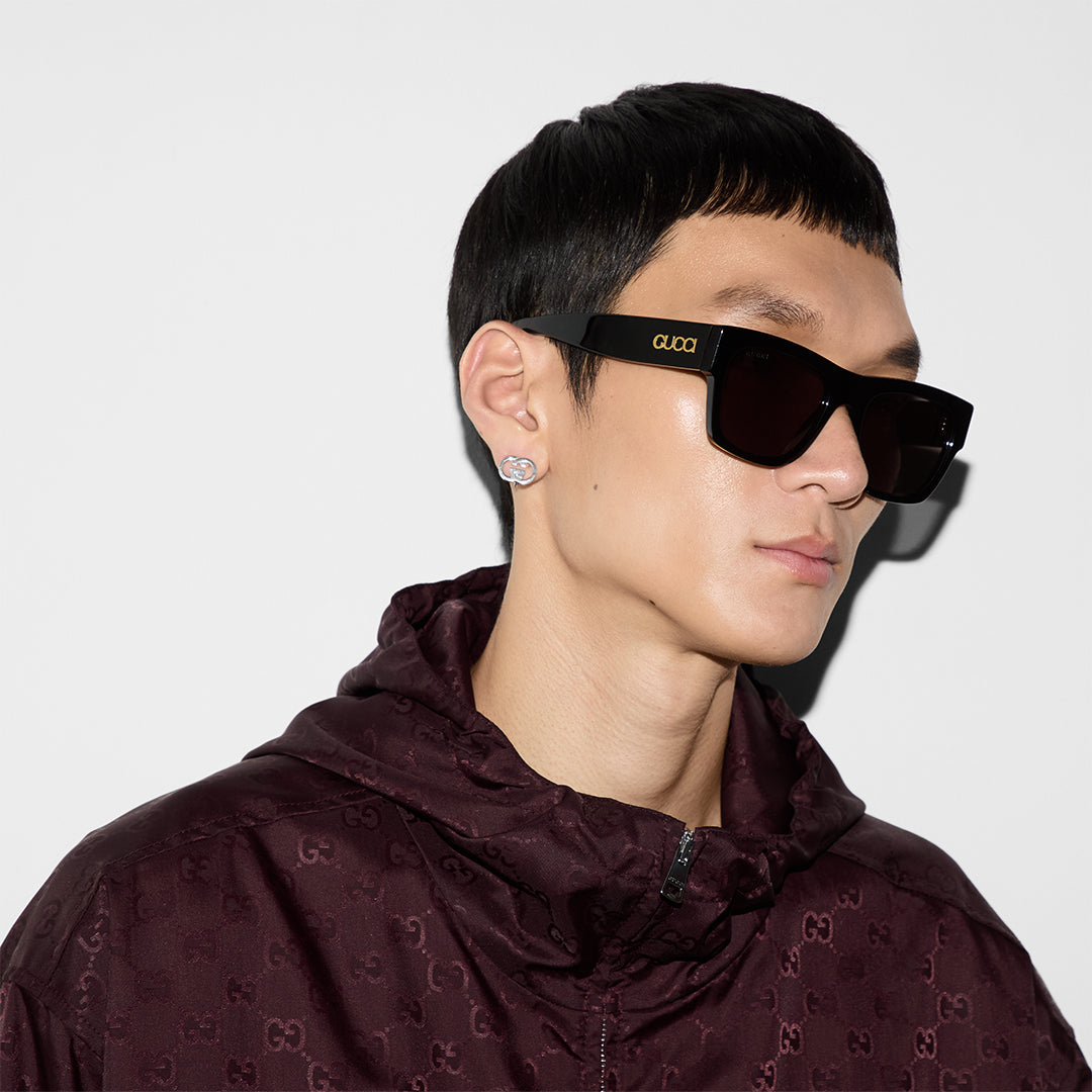 A person wearing black square sunglasses with "GUCCI" on the side, a maroon Gucci-patterned jacket, and GUCCI Fine Jewellery Interlocking Sterling Silver Stud Earrings, looking to the right against a plain white background.