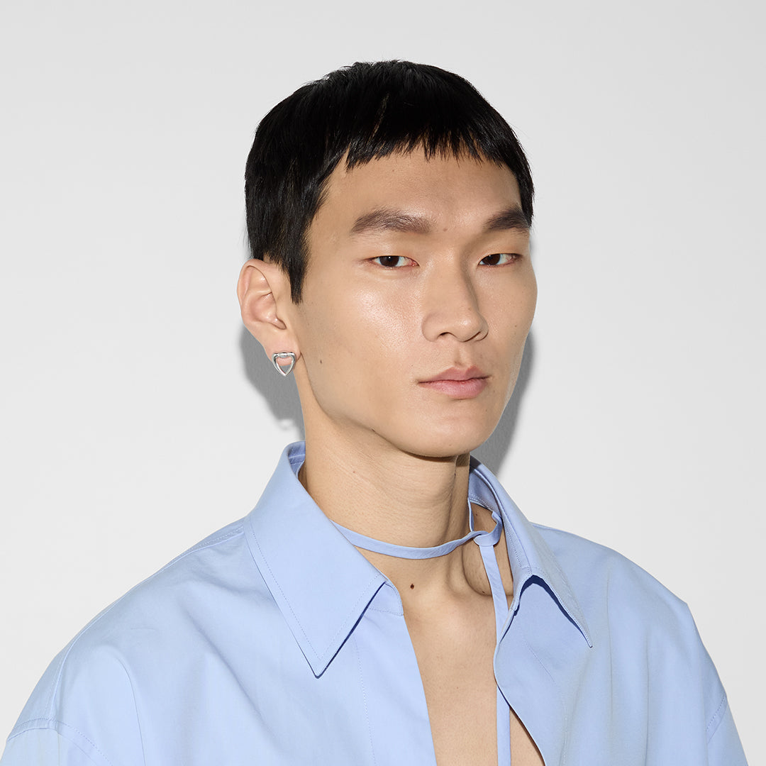 A person with short black hair is wearing a light blue collared shirt with a tie and Gucci Sterling Silver Heart Stud Earrings by GUCCI Fine Jewellery, standing against a white background.