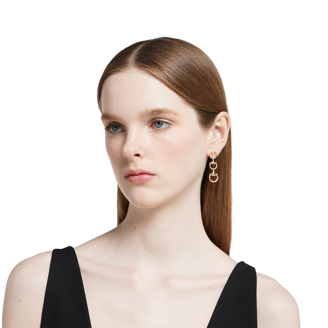 A woman with straight brown hair wears a sleeveless black top and GUCCI Fine Jewellery Horsebit 18K Yellow Gold Drop Earrings, looking slightly to the side against a white background.