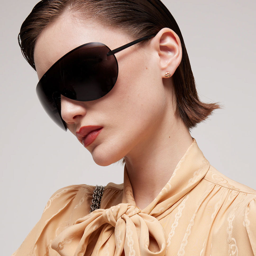 A woman with short brown hair wears oversized black sunglasses, Gucci Interlocking G 18K Yellow Gold Stud Earrings from GUCCI Fine Jewellery, and a beige blouse with a bow at the collar as she looks to the side against a plain background.