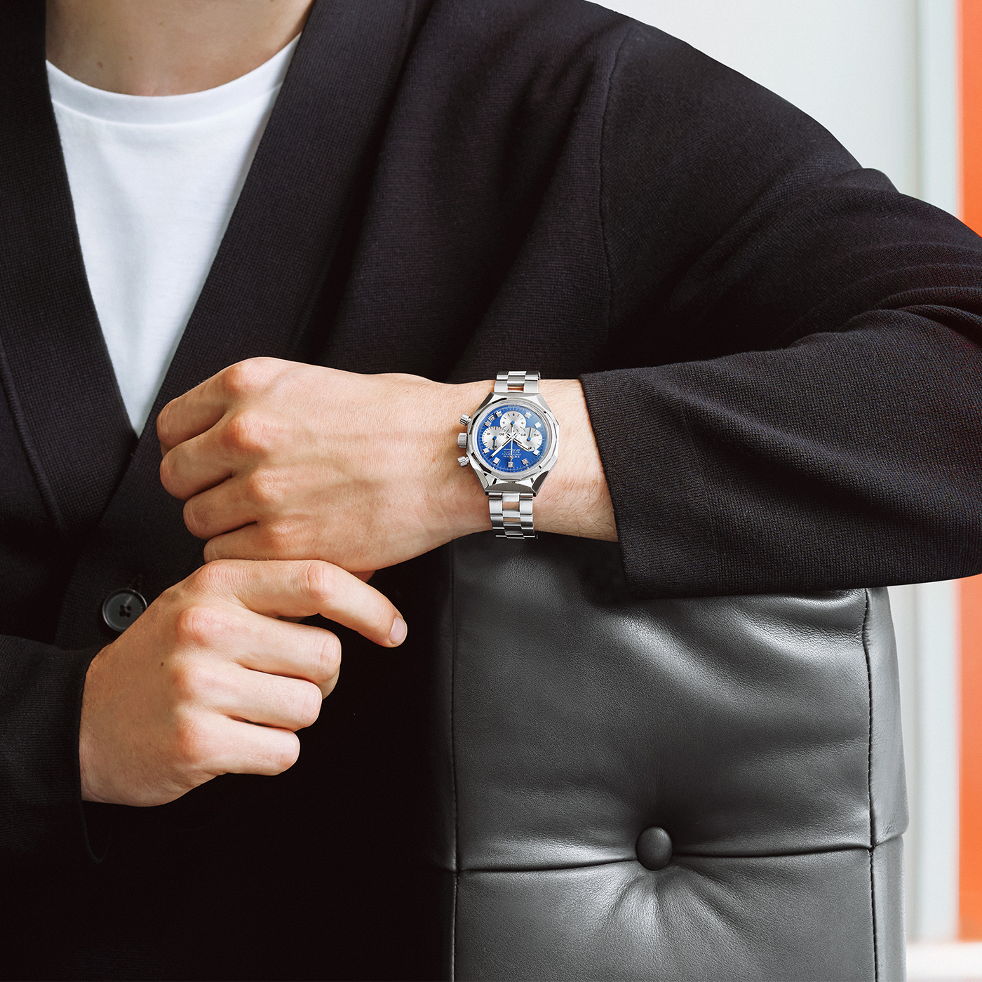 A person in a black jacket and white shirt sits with their arm on a gray leather chair, wearing the Zenith Defy Chronograph USM - Genetian Blue, a limited edition silver wristwatch by Zenith, on their left wrist.